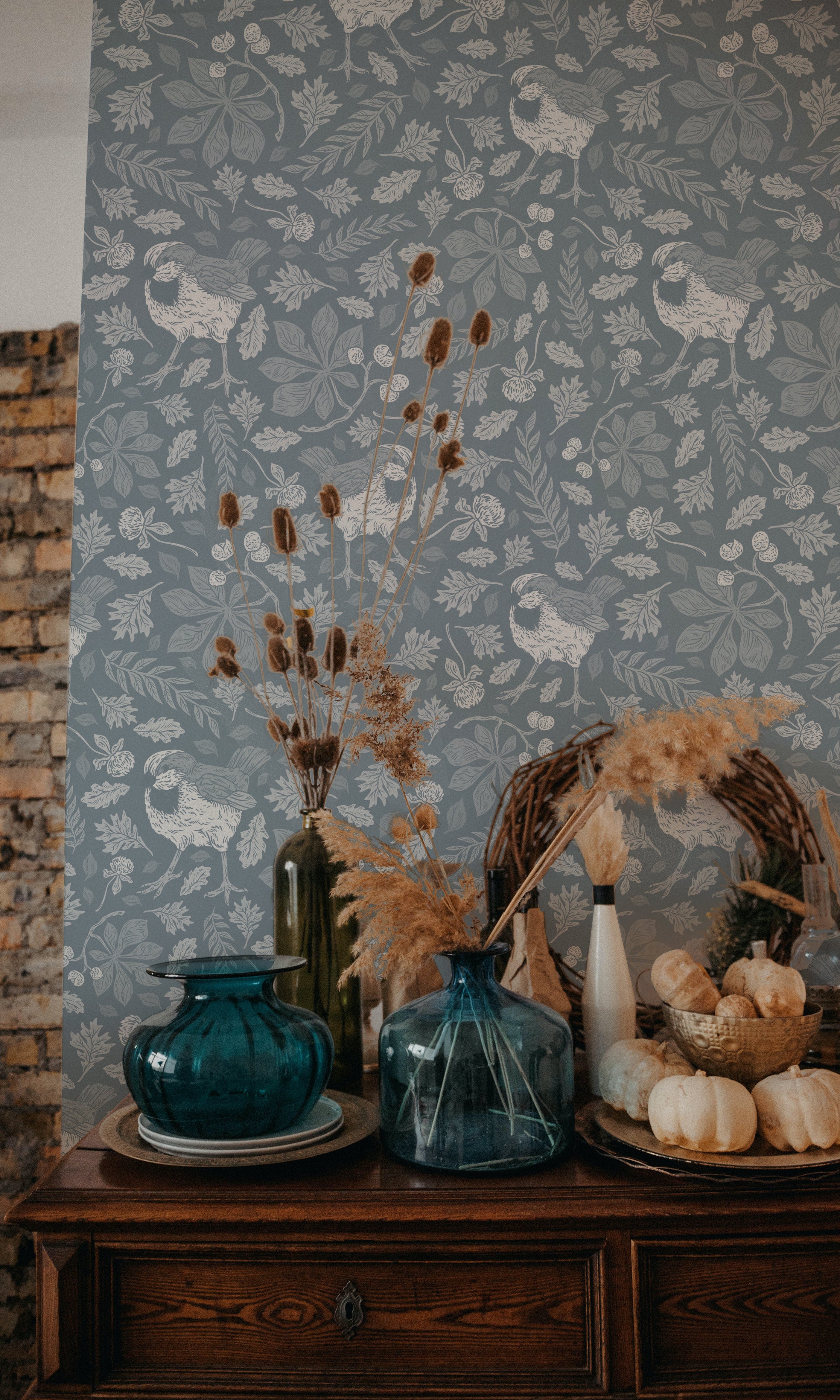 A rustic interior scene featuring a wall covered with vintage-style sparrow wallpaper in a cool grey color. The wallpaper is illustrated with white sparrows and delicate foliage. In the foreground, a dark wooden sideboard displays blue glass vases, dried flowers, and white pumpkins.