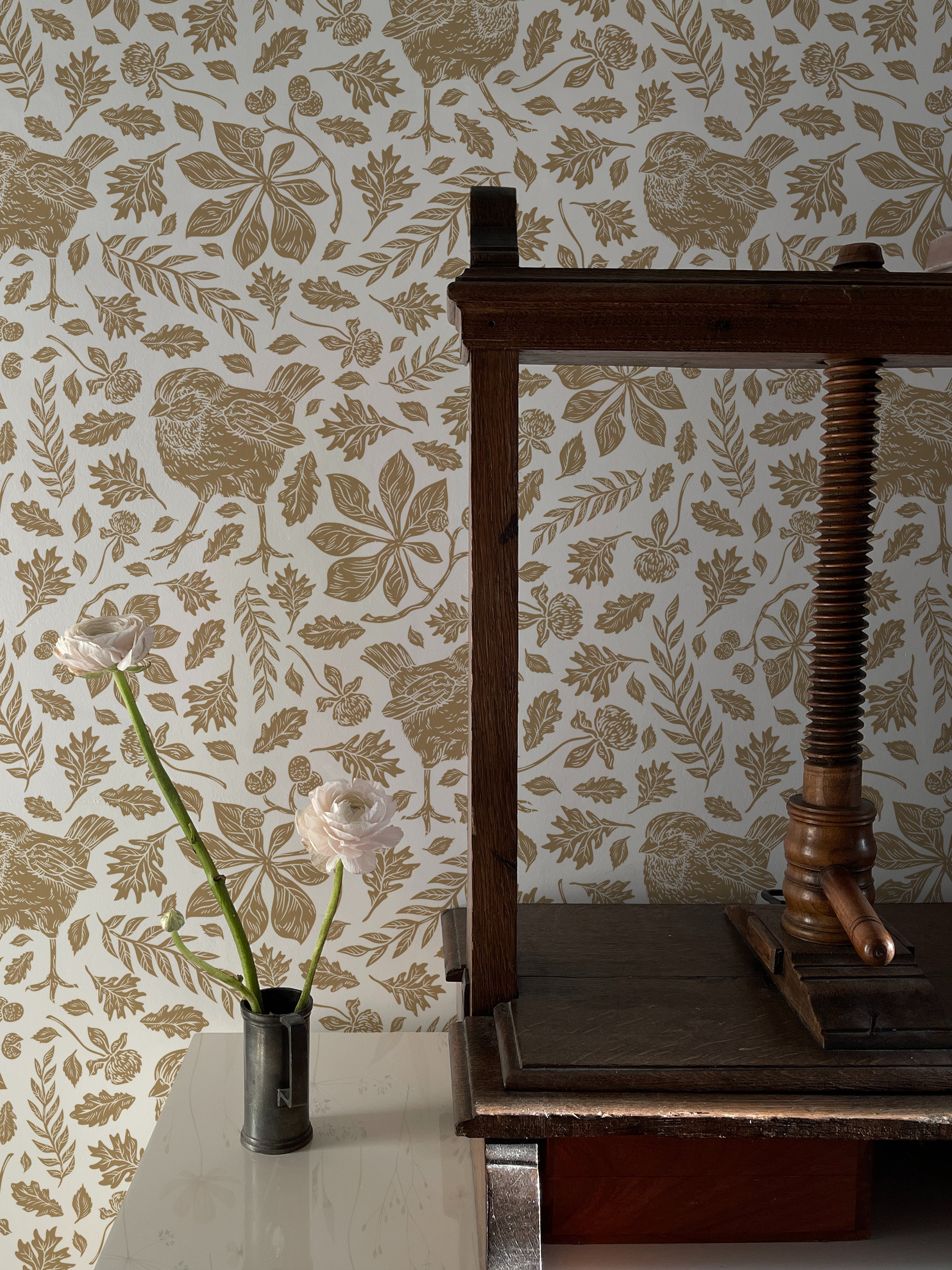 A detailed view of a vintage-style wallpaper featuring golden sparrows and an assortment of leaves and floral patterns on a cream background. An antique wooden spindle stands against the wall, with two light pink flowers in a small metal vase placed on a marble tabletop.