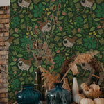 A vibrant green version of the vintage sparrow wallpaper decorates a wall, creating a bold backdrop to a rustic scene with a wooden sideboard, blue glass vases, dried plants, and white pumpkins. The wallpaper features brown sparrows amidst green foliage with small yellow and orange details.