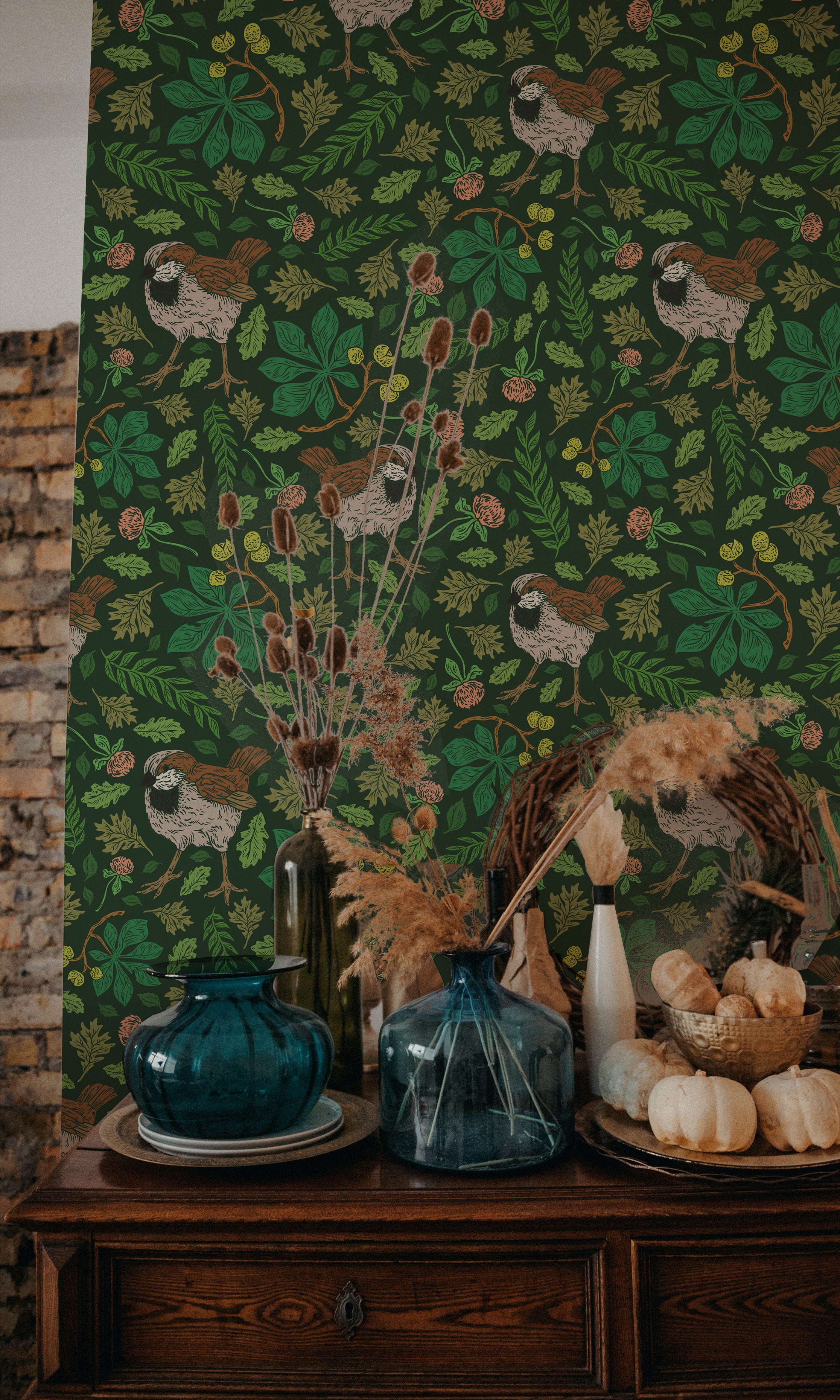 A vibrant green version of the vintage sparrow wallpaper decorates a wall, creating a bold backdrop to a rustic scene with a wooden sideboard, blue glass vases, dried plants, and white pumpkins. The wallpaper features brown sparrows amidst green foliage with small yellow and orange details.