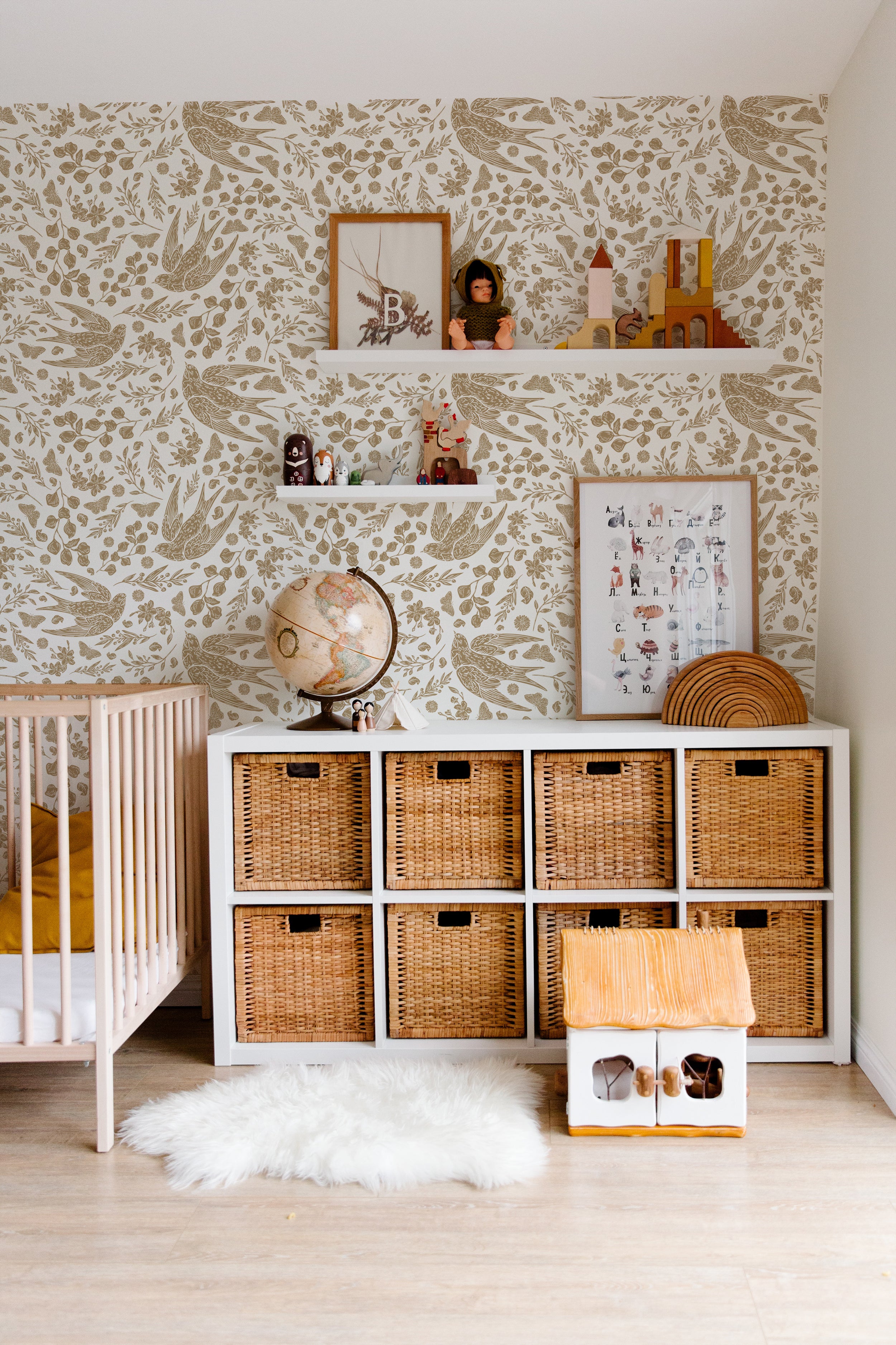 A cozy nursery room corner featuring Vintage Swift Wallpaper with elegant golden bird and foliage illustrations on a cream background. The room includes a crib, a shelf filled with children's toys and books, and a white storage unit with wicker baskets