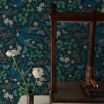 Elegant interior with Vintage Swift Wallpaper featuring dark teal background adorned with intricate illustrations of swift birds in flight, interspersed with delicate foliage and golden accents. A rustic wooden spindle and white roses in a metal vase enhance the vintage charm.