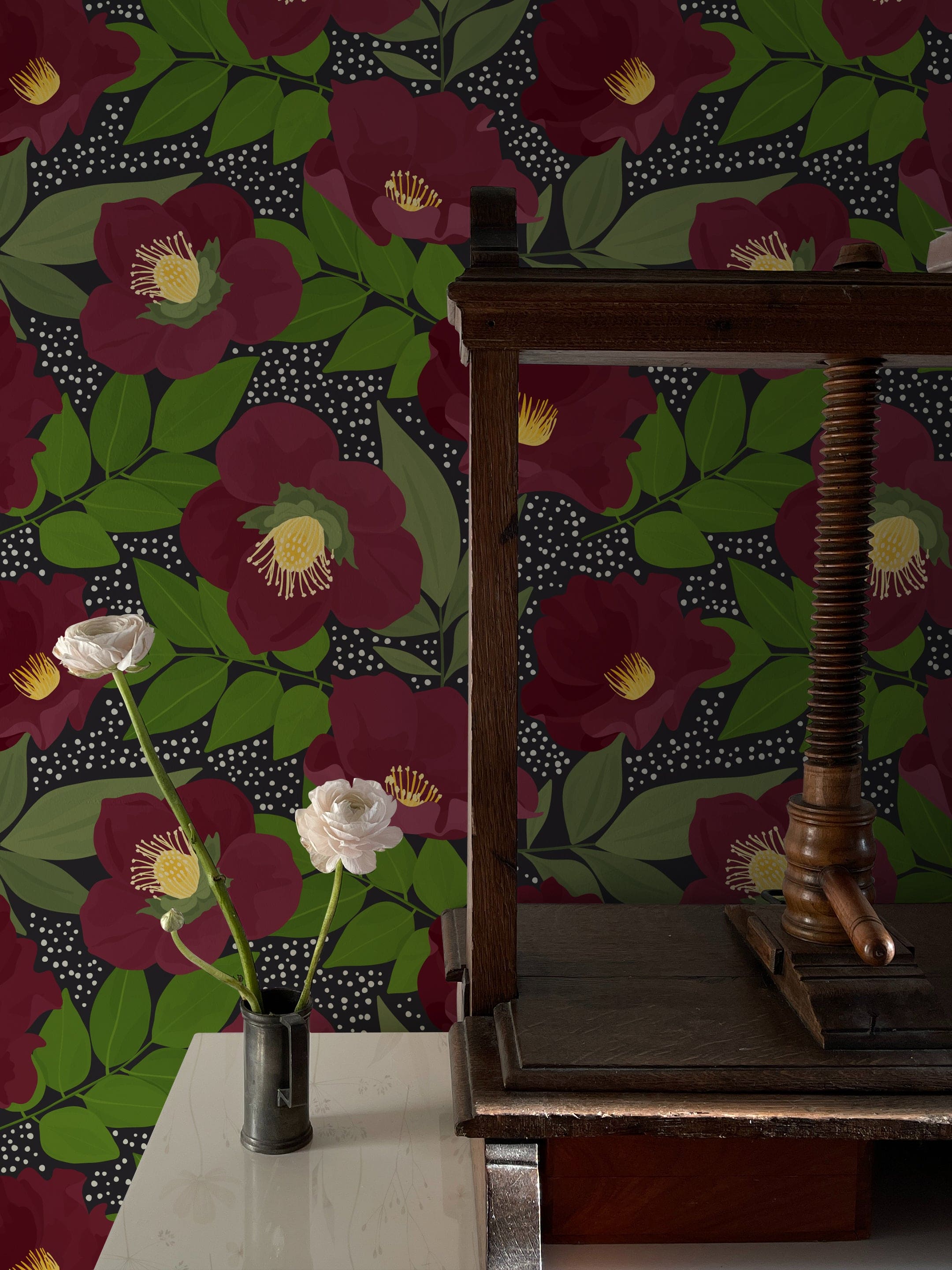 A sophisticated corner of a room showcasing the Merlot Floral Wallpaper, with its bold, merlot-colored blooms and vibrant green leaves on a dark, dotted background. The floral design is complemented by a vintage wooden table and a simple white rose in a black vase.