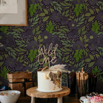 A festive setting with the Martinique Floral Wallpaper enhancing the background of a dessert table featuring a birthday cake and snacks. The wallpaper's rich purple florals and lush green leaves add a vibrant and celebratory atmosphere to the scene.