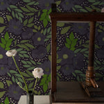 A stylish interior space with the Martinique Floral Wallpaper, showcasing a dark and moody floral pattern with large purple blooms and vibrant green foliage. A rustic wooden table holds a single white rose in a small vase, contrasting elegantly against the detailed wallpaper.