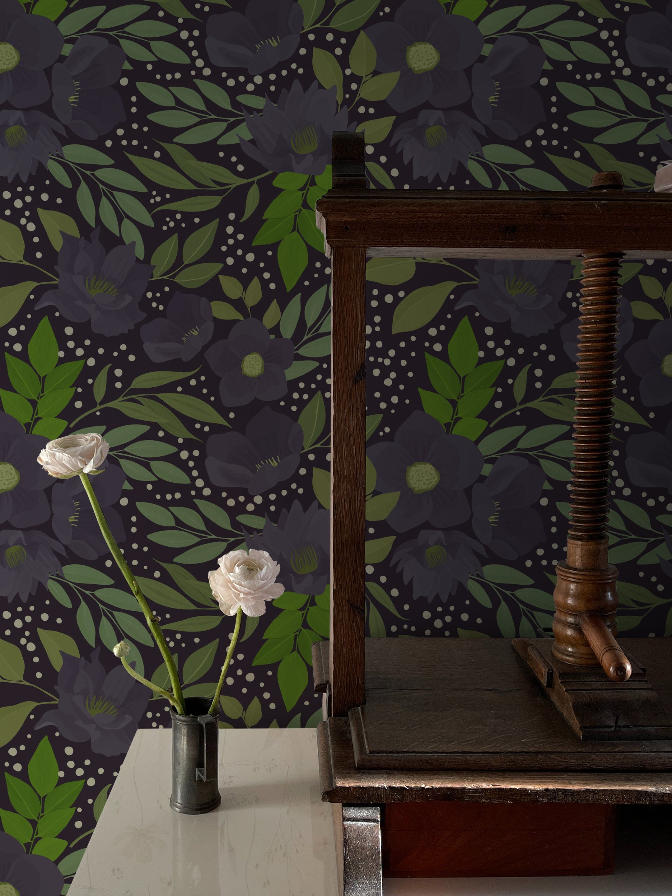 A stylish interior space with the Martinique Floral Wallpaper, showcasing a dark and moody floral pattern with large purple blooms and vibrant green foliage. A rustic wooden table holds a single white rose in a small vase, contrasting elegantly against the detailed wallpaper.