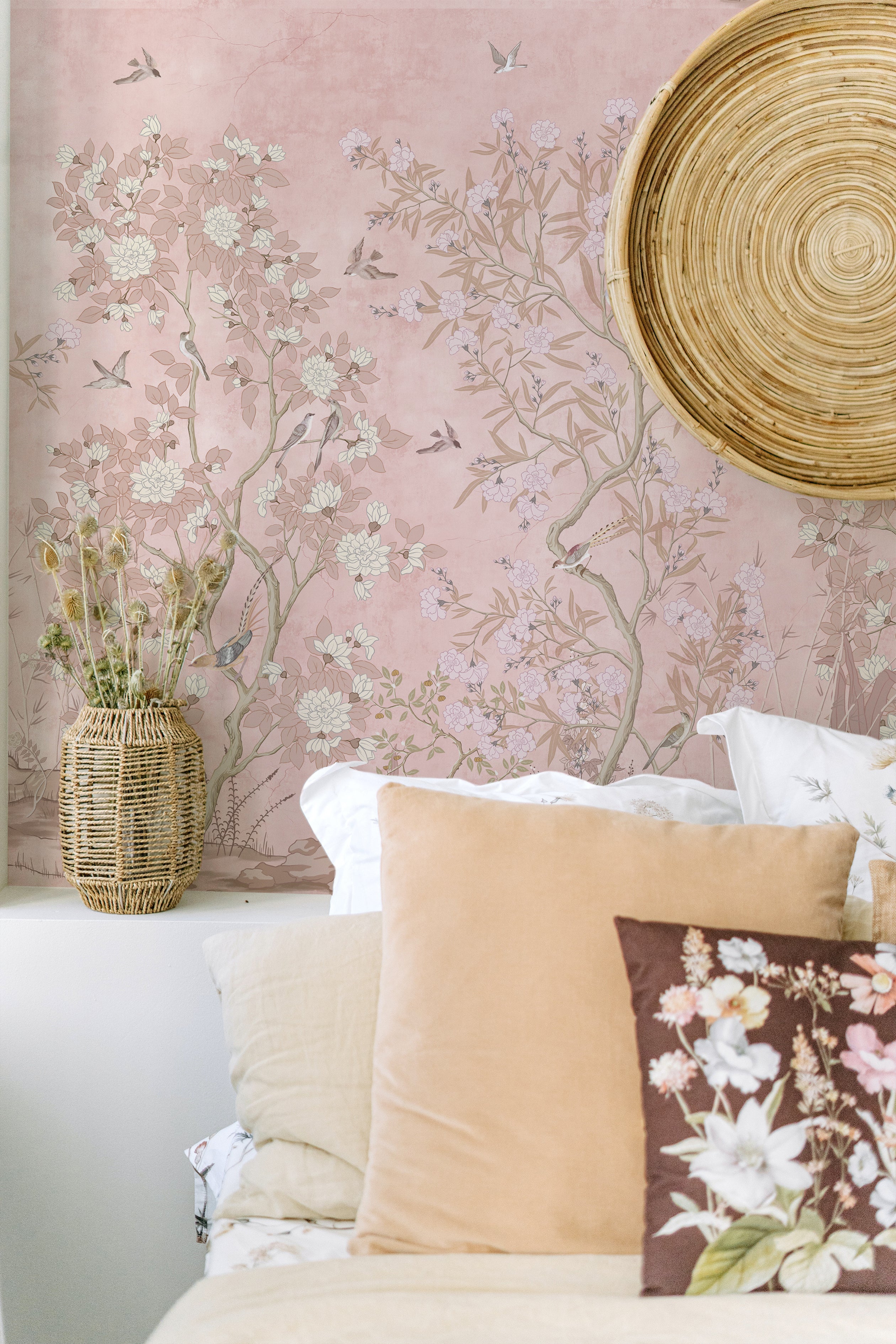 A close-up of chinoiserie wallpaper behind a bedroom scene, showing intricate details of the floral and avian pattern in muted colors on a blush pink background, complemented by a natural wicker headboard and accents like a large straw wall disc, fresh flowers in a basket, and soft-colored pillows