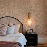 A bedroom featuring chinoiserie wallpaper with a delicate design of trees, blooming flowers, and birds in soft colors on a pale pink background, providing a tranquil backdrop to a bed with a dark, arched headboard, layered with textured linens in shades of white and dusty rose