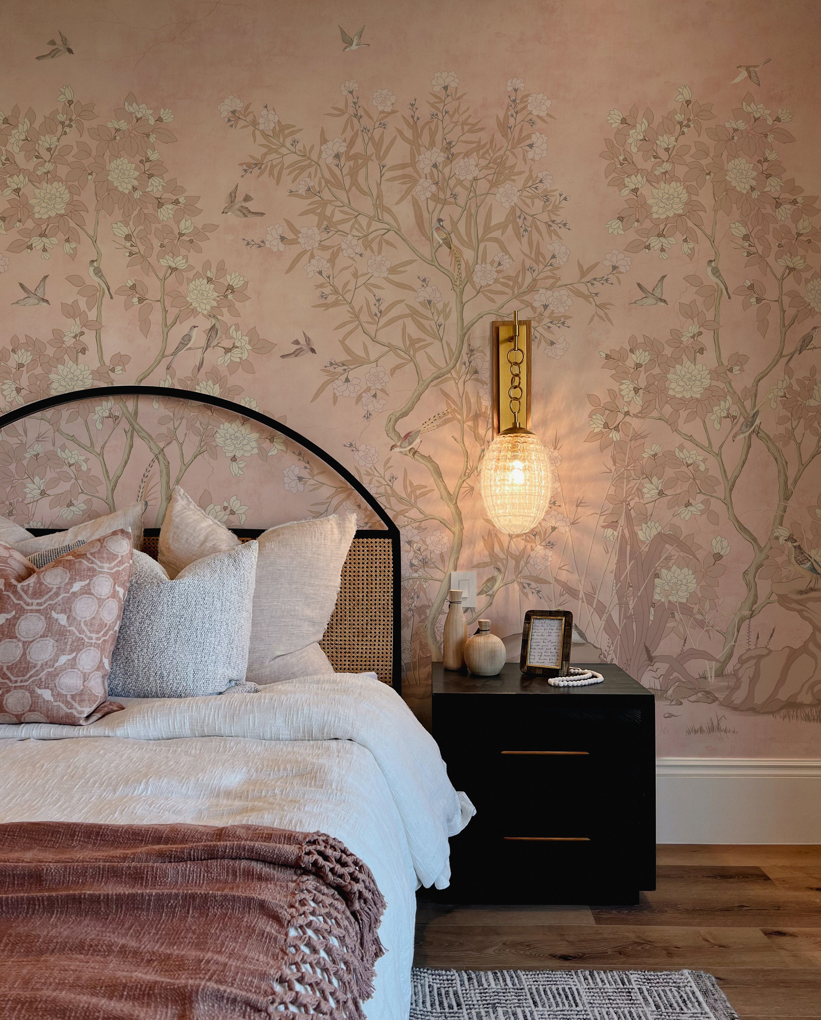 A bedroom featuring chinoiserie wallpaper with a delicate design of trees, blooming flowers, and birds in soft colors on a pale pink background, providing a tranquil backdrop to a bed with a dark, arched headboard, layered with textured linens in shades of white and dusty rose