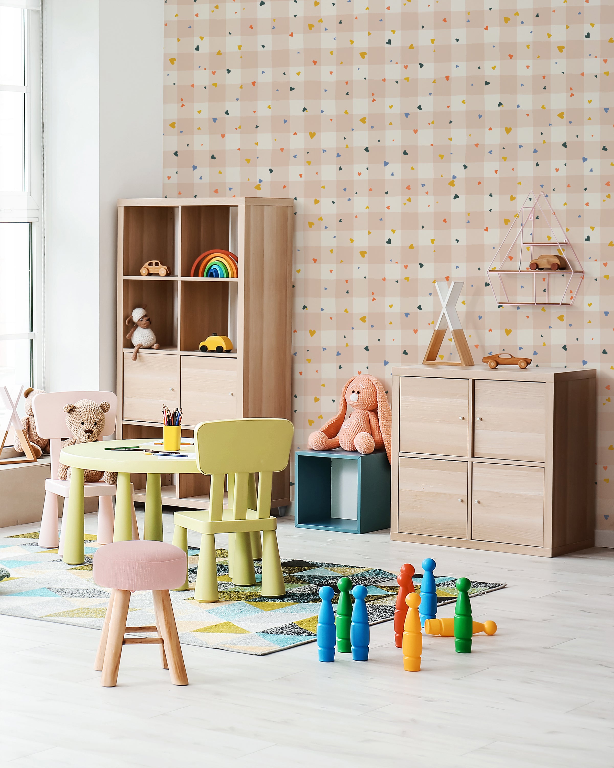 A children's playroom with walls covered in a cheerful pink wallpaper adorned with a colorful confetti-like pattern of triangles, paired with wooden furniture and bright toys