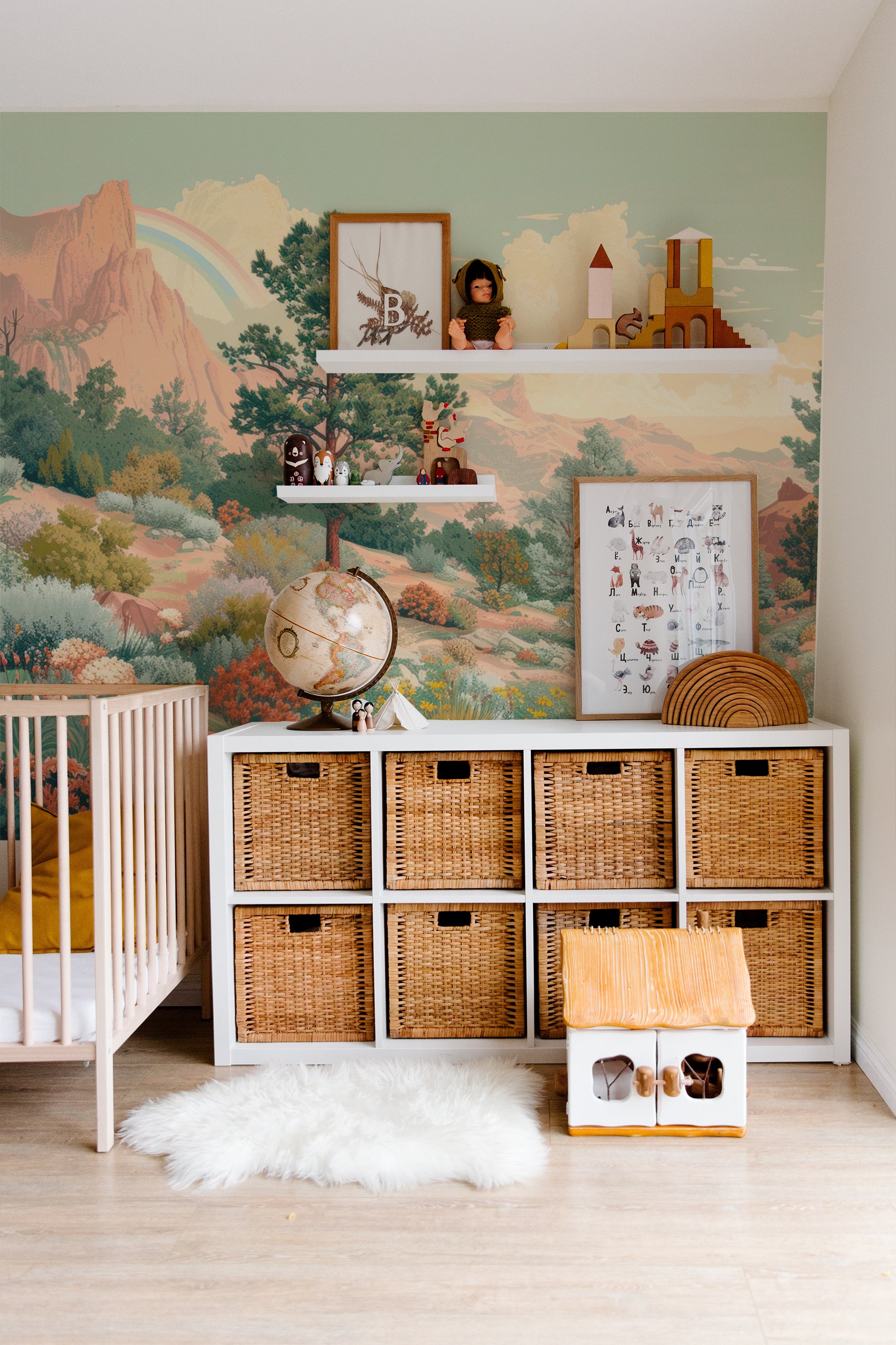 A nursery room decorated with a desert-themed wallpaper showing a mountainous landscape under a bright sky. The room includes a crib, a wicker basket storage unit, and decorative items that complement the earthy tones of the mural.