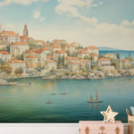 "Wallpaper mural of Dubrovnik coastline in a nursery, decorated with a white chest of drawers, plush whale, and toys.