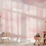 A playful children's room with a white table and chair set, adorned with a wooden shelf holding pink flowers. The wall is decorated with Pink Cloud Mural wallpaper, featuring fluffy pink and white clouds that add a dreamy touch to the space