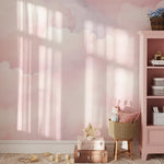 A cozy children's corner with a wicker basket, plush toys, and a pink bookshelf. The wall behind is covered with Pink Cloud Mural wallpaper, displaying a serene pattern of pink and white clouds, creating a calming and imaginative environment.