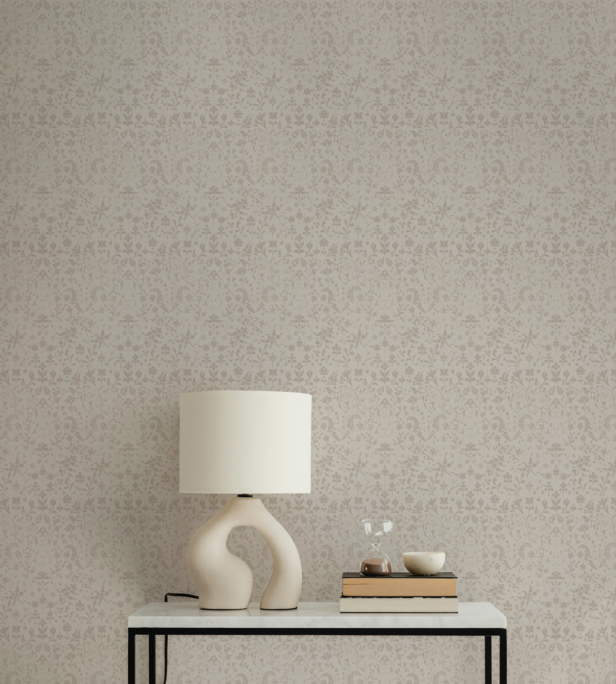 A chic console table with a sculptural white lamp, a stack of books, and decorative items. The wall behind features the Frontenac Wallpaper, presenting a refined and intricate pattern in light taupe, perfect for adding an elegant touch to any space.