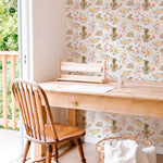 A cozy crafting area featuring the Garden Bunnies Wallpaper, which displays playful rabbits nestled in baskets among soft floral designs. The scene includes a sturdy wooden chair and table, creating a warm, inviting workspace.