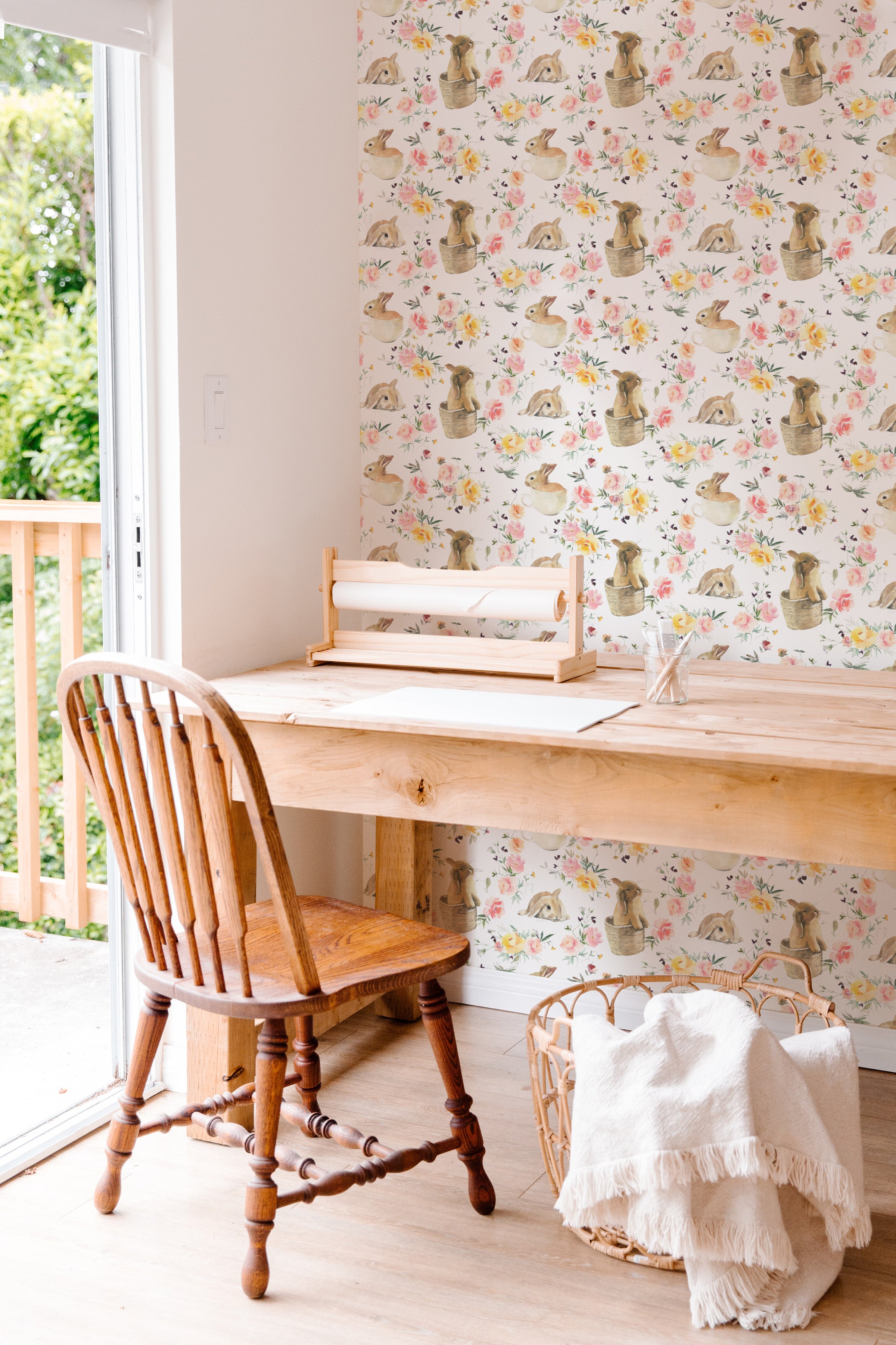 A cozy crafting area featuring the Garden Bunnies Wallpaper, which displays playful rabbits nestled in baskets among soft floral designs. The scene includes a sturdy wooden chair and table, creating a warm, inviting workspace.