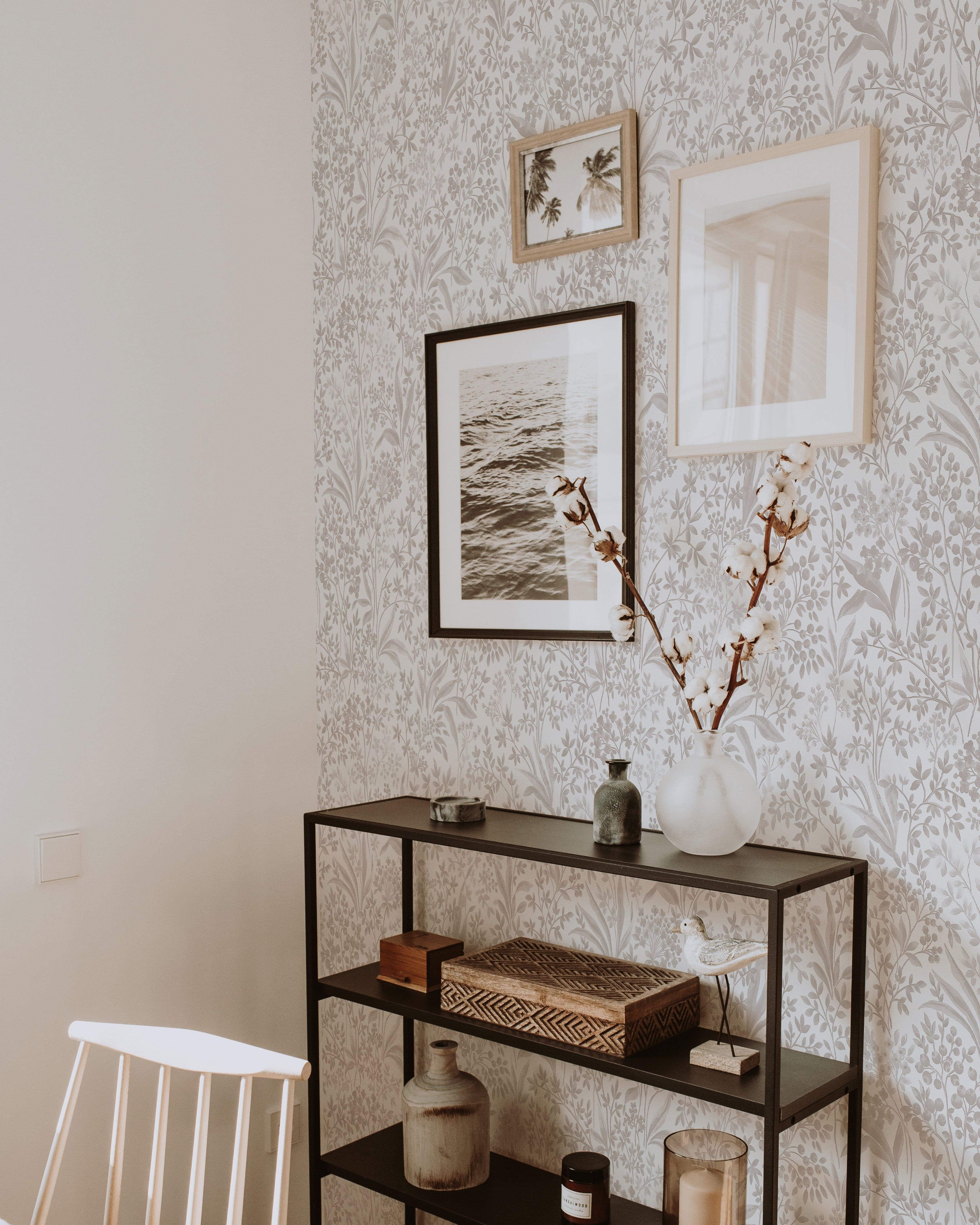 A modern display area enhanced by the Harmony in Bloom Wallpaper, showcasing a dense floral design in soft gray and beige tones. The wall is accessorized with framed photographs and a minimalist black metal shelf with decorative items, blending contemporary design with classic elegance.