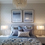 A coastal-inspired bedroom showcasing twin beds with blue ocean-themed bedding and pillows. The walls are adorned with the same light gray scallop shell wallpaper as in the previous images. Above the bed hang white capiz shell chandeliers, and framed oceanic art completes the serene maritime decor