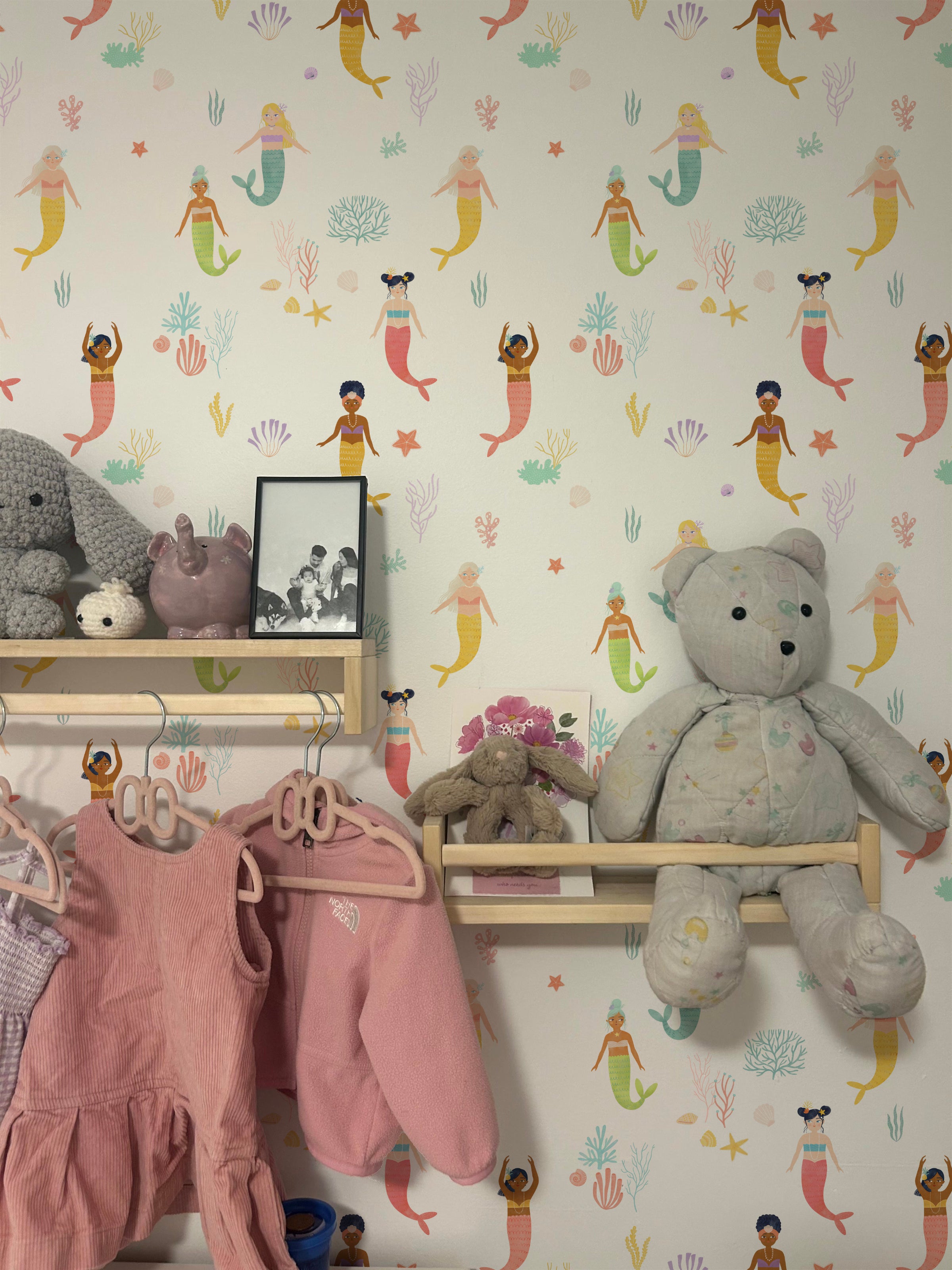 A charming children's room decorated with Little Mermaids Wallpaper, featuring playful mermaids in a variety of poses and colors, accompanied by marine flora and fauna on a light background. The scene includes a display shelf with plush toys and children's clothes, creating a whimsical and joyful space.