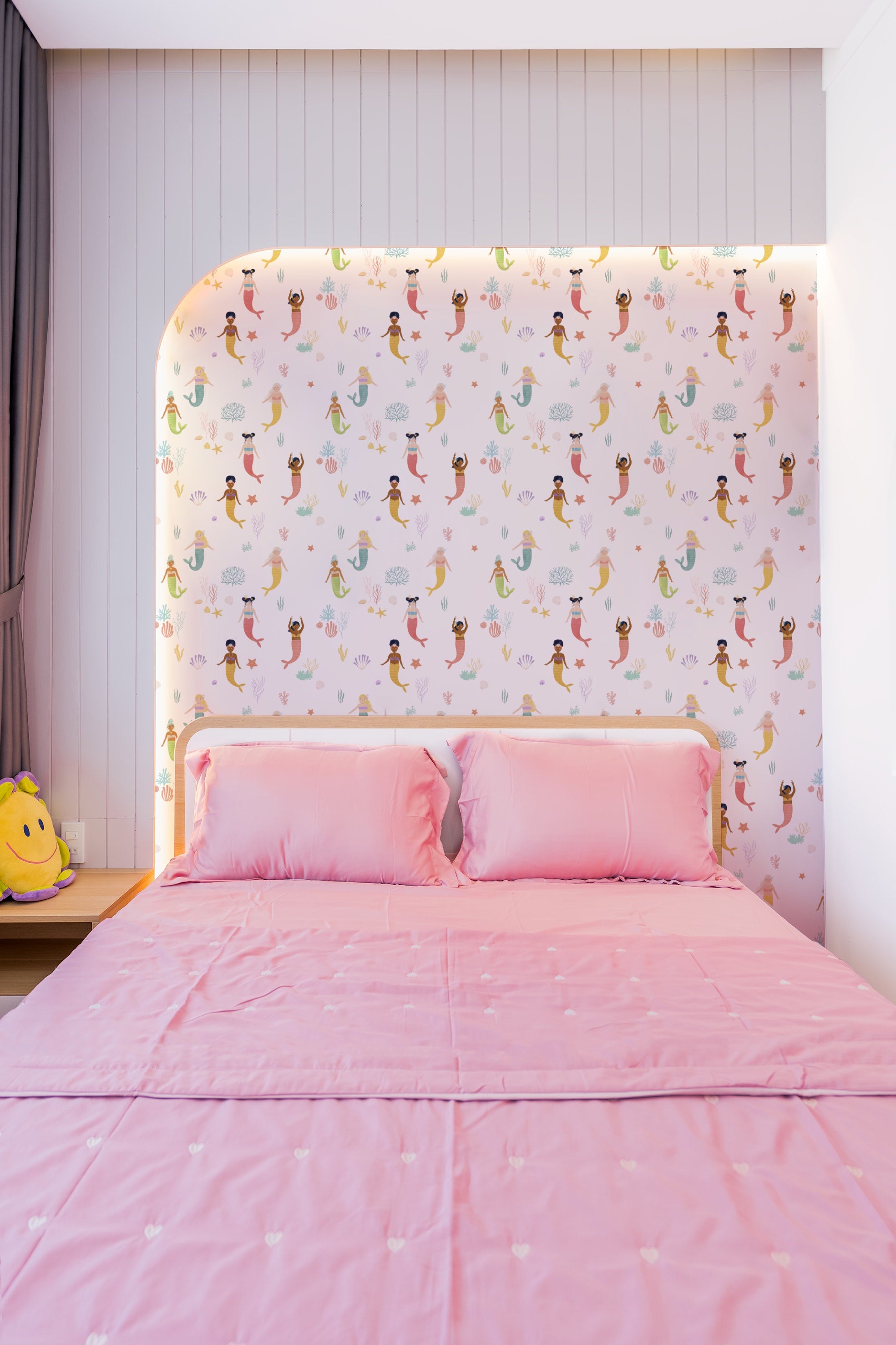 A cozy bedroom setting with Little Mermaids Wallpaper providing a lively and imaginative backdrop. The bed is dressed in pink linens, complementing the colorful mermaids and enhancing the room’s playful and magical atmosphere.