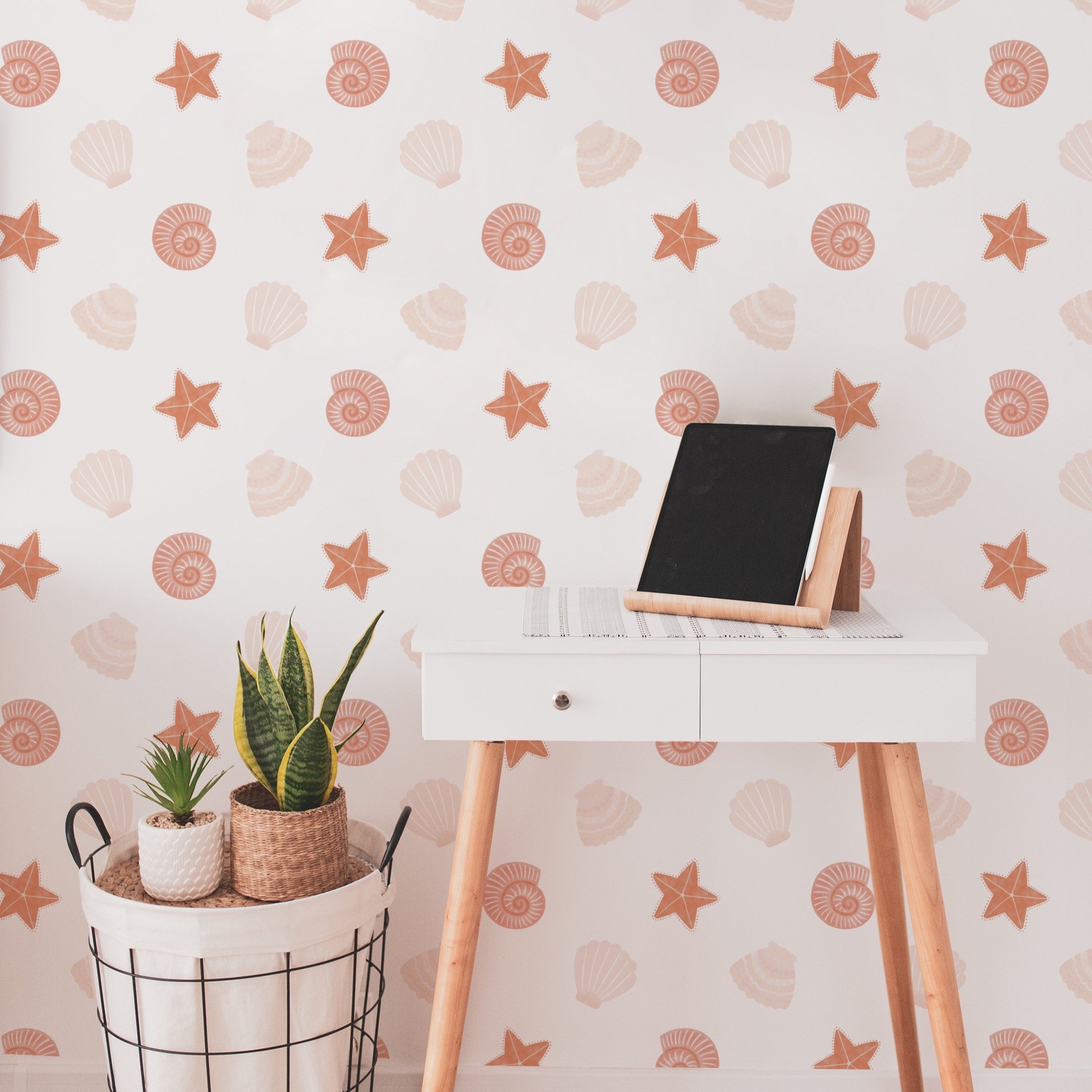 A chic home office setup with Mermaid Shells Wallpaper, creating a bright and inviting workspace. The wall features a pattern of delicate seashells and starfish, complemented by a modern white desk and green houseplants.