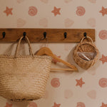 A functional wall space featuring Mermaid Shells Wallpaper adorned with seashell and starfish patterns in soft orange tones on a beige background. Rustic wooden hooks hold a woven basket and a wooden hanger, enhancing the nautical theme.