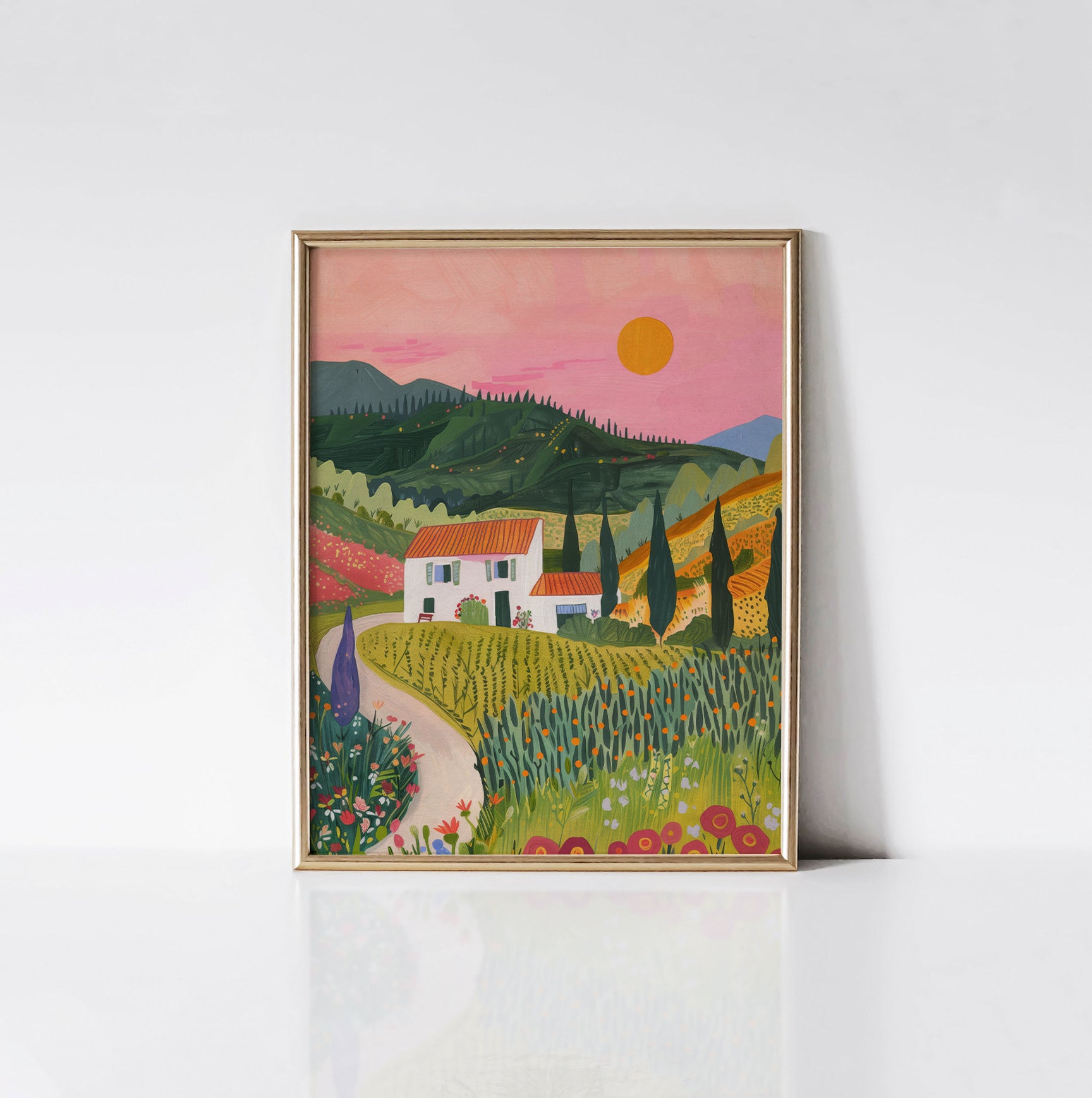 The 'Sunny Countryside Retreat' art print displayed in a sleek gold frame, showcasing a white cottage amidst green fields and blooming flowers with a pink sunset in the background.