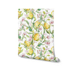 A roll of Citrus Blossom Wallpaper partially unrolled to display its charming pattern of yellow lemons and pink blossoms on a background of lush greenery. This image emphasizes the wallpaper’s ability to brighten and enrich any interior with its vibrant and cheerful design
