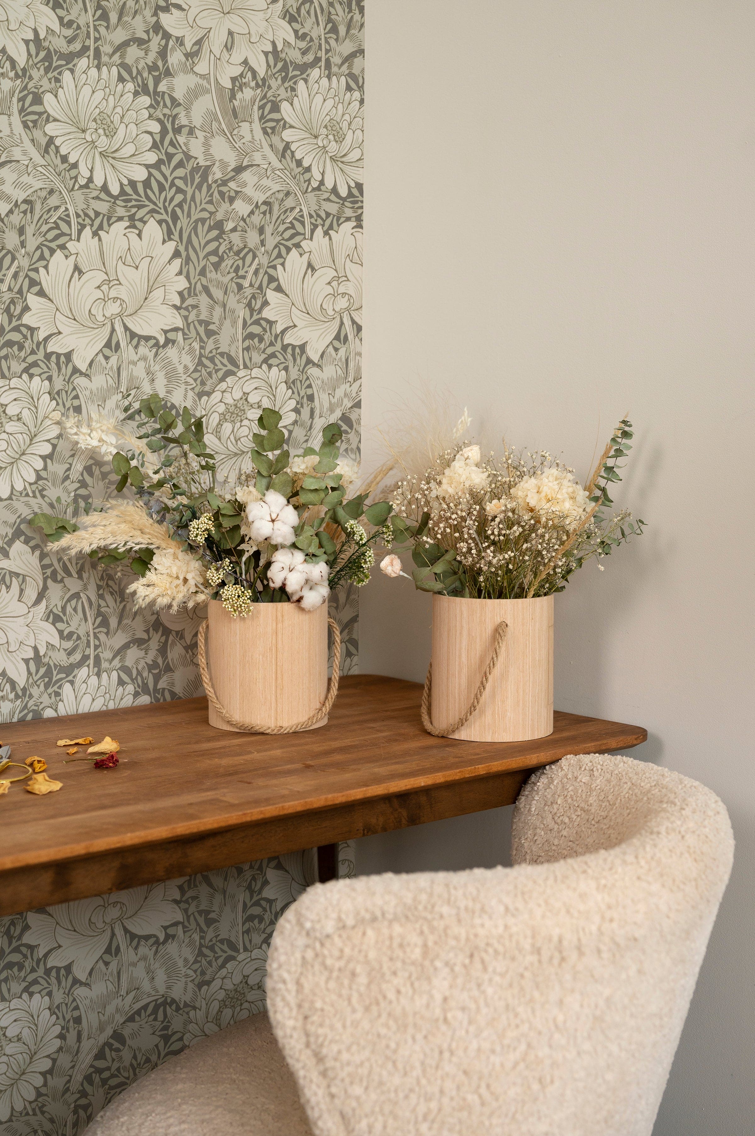 A cozy interior corner featuring the Entwined Elegance Wallpaper with a vintage floral pattern in grayscale. Two natural wood vases with dried white flowers sit atop a wooden shelf, adding a touch of rustic charm against the sophisticated backdrop.