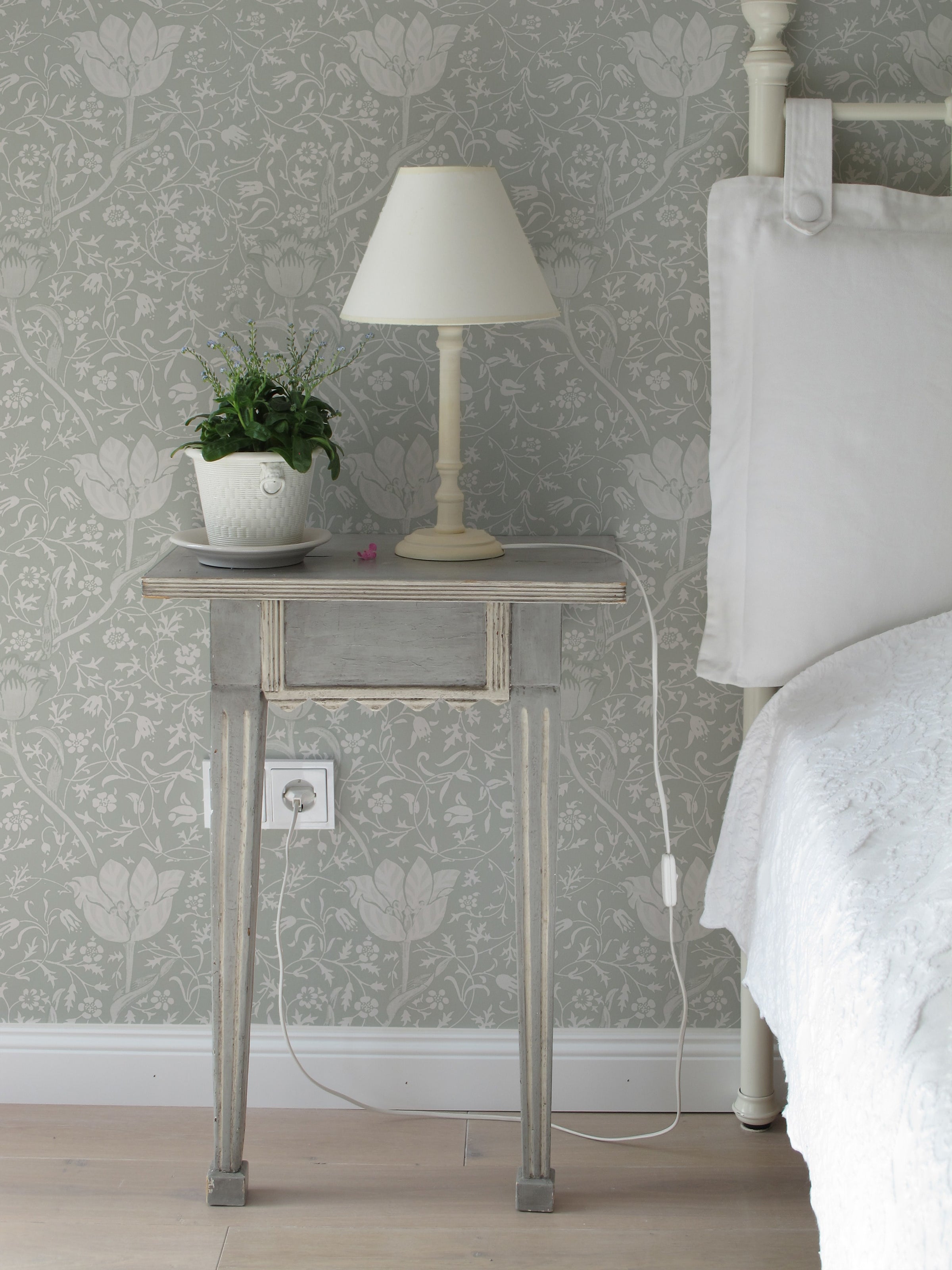 A vintage-inspired bedroom corner showcasing Fiona Floral Wallpaper in Light Sage. The wall features a detailed botanical print with large floral motifs in white over a soft sage background. A rustic wooden side table holds a classic lamp and a potted plant, enhancing the old-world charm.