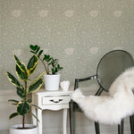 A chic interior setup displaying Fiona Floral Wallpaper in Light Sage. The wall adorned with detailed floral patterns creates a refined backdrop for a modern white side table and a transparent chair draped with a white fur throw. A potted fiddle-leaf fig tree adds a natural touch.