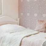 A cozy bedroom corner with a wall covered in Fiona Floral Wallpaper in soft pink with detailed mauve flowers. The room features a white bed with pale pink bedding and a plush gray blanket, creating a warm and inviting atmosphere.