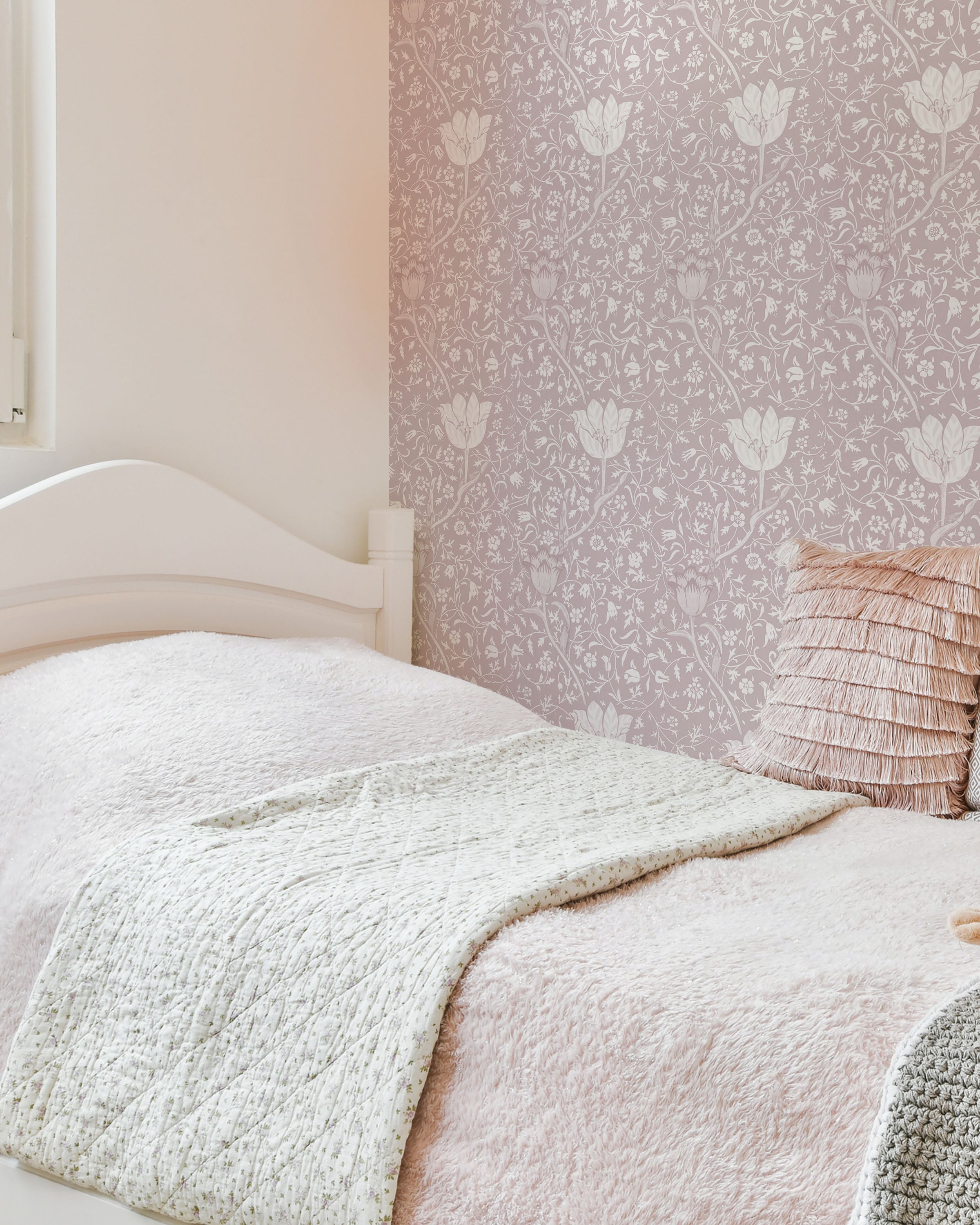 A cozy bedroom corner with a wall covered in Fiona Floral Wallpaper in soft pink with detailed mauve flowers. The room features a white bed with pale pink bedding and a plush gray blanket, creating a warm and inviting atmosphere.
