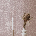 A styled scene showcasing Fiona Floral Wallpaper, featuring a detailed floral pattern in mauve on a soft pink background. The setup includes a modern candle holder and a textured ceramic vase with dried botanicals, creating a serene and stylish space