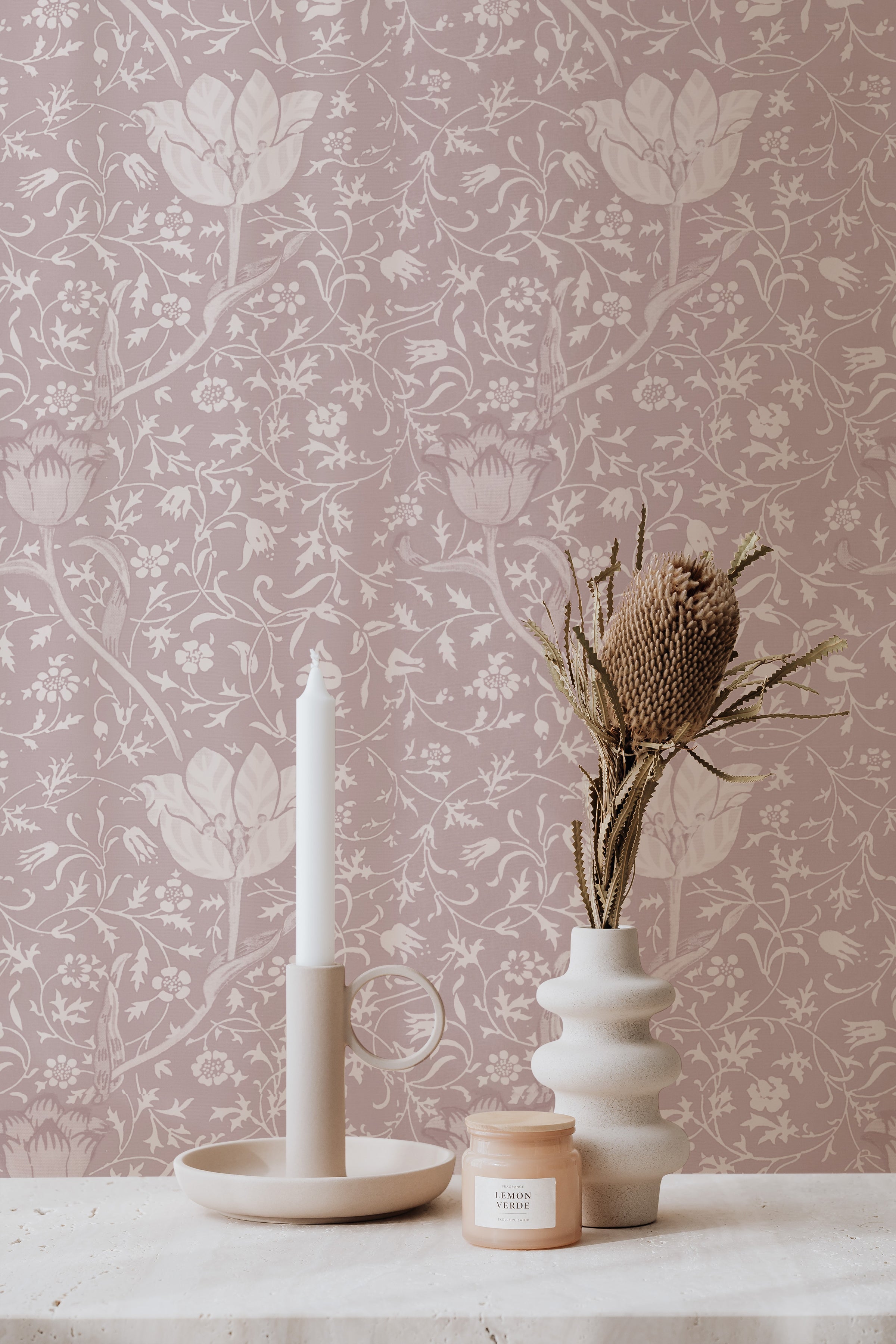 A styled scene showcasing Fiona Floral Wallpaper, featuring a detailed floral pattern in mauve on a soft pink background. The setup includes a modern candle holder and a textured ceramic vase with dried botanicals, creating a serene and stylish space