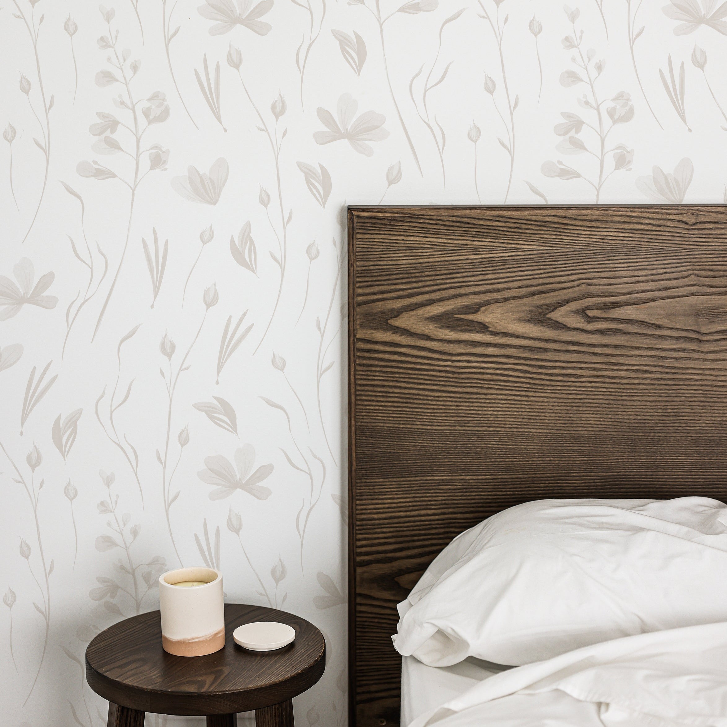A bedroom scene with the wall featuring warm grey watercolor floral wallpaper, adding a tranquil and elegant atmosphere, alongside a dark wooden bed frame and a small round side table with a ceramic cup.