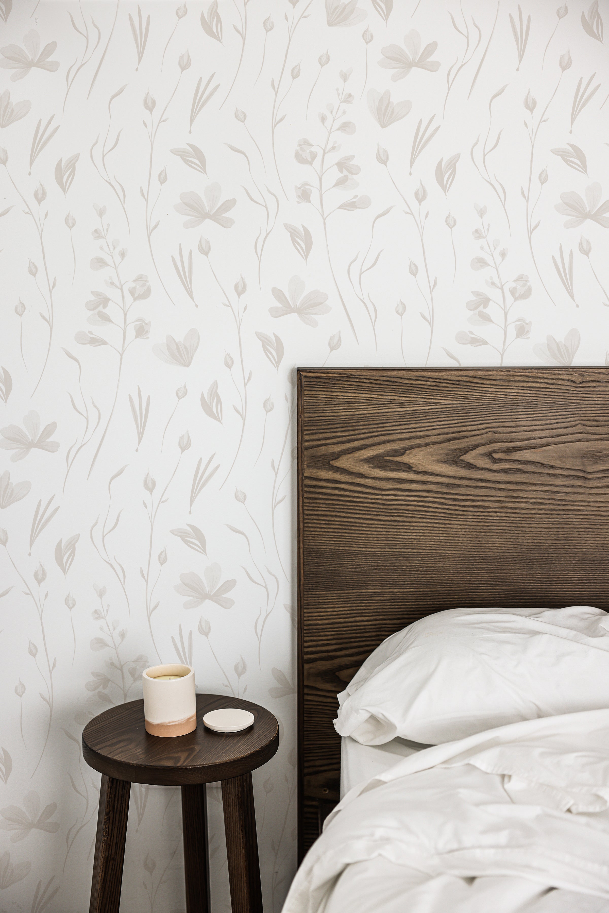 A bedroom scene with the wall featuring warm grey watercolor floral wallpaper, adding a tranquil and elegant atmosphere, alongside a dark wooden bed frame and a small round side table with a ceramic cup.