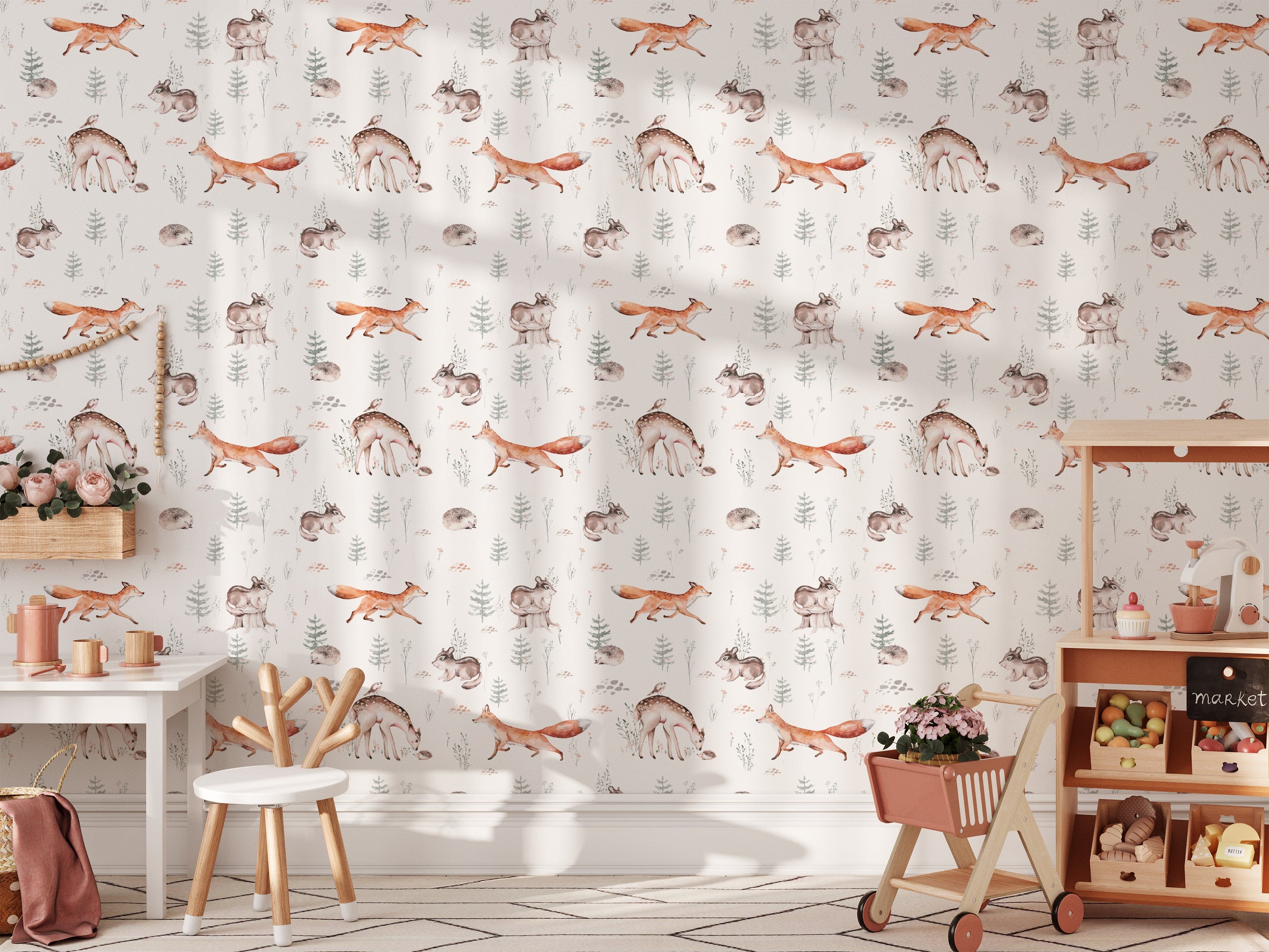 A delightful children's playroom decorated with Woodland Wonder Wallpaper featuring charming illustrations of forest animals such as foxes, deer, rabbits, and hedgehogs amidst trees and foliage. The scene is complemented by a kid-friendly desk and chair set, a toy cart, and vibrant room decor.