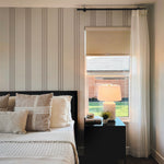 A cozy bedroom corner where the Fabric 5F Wallpaper provides a calming backdrop with its subtle ticking stripe pattern in gray on a neutral field. The wallpaper gives the illusion of fabric on the walls, complementing the texture of the bedding and the warm glow from a bedside lamp, contributing to a warm and inviting atmosphere.