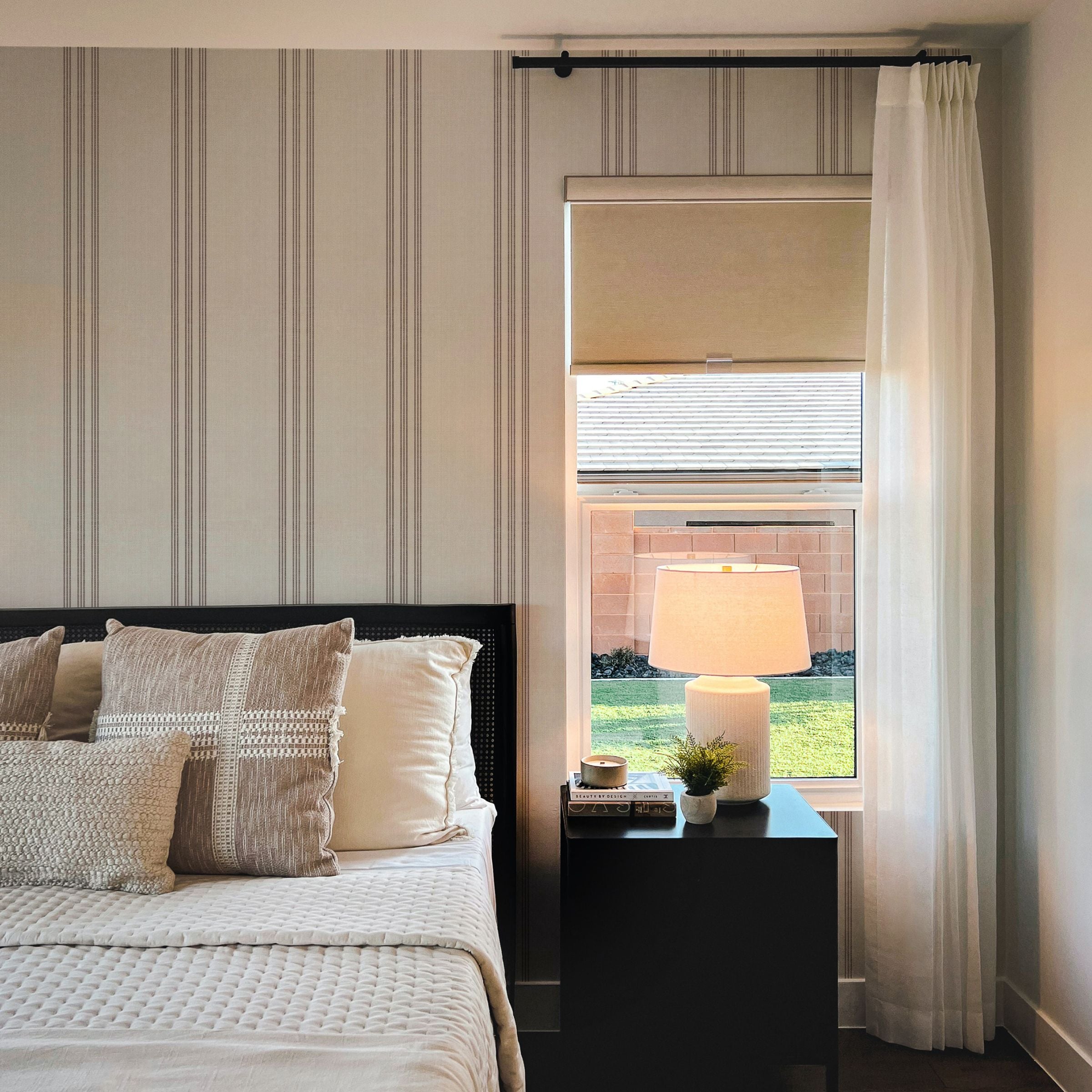 A cozy bedroom corner where the Fabric 5F Wallpaper provides a calming backdrop with its subtle ticking stripe pattern in gray on a neutral field. The wallpaper gives the illusion of fabric on the walls, complementing the texture of the bedding and the warm glow from a bedside lamp, contributing to a warm and inviting atmosphere.