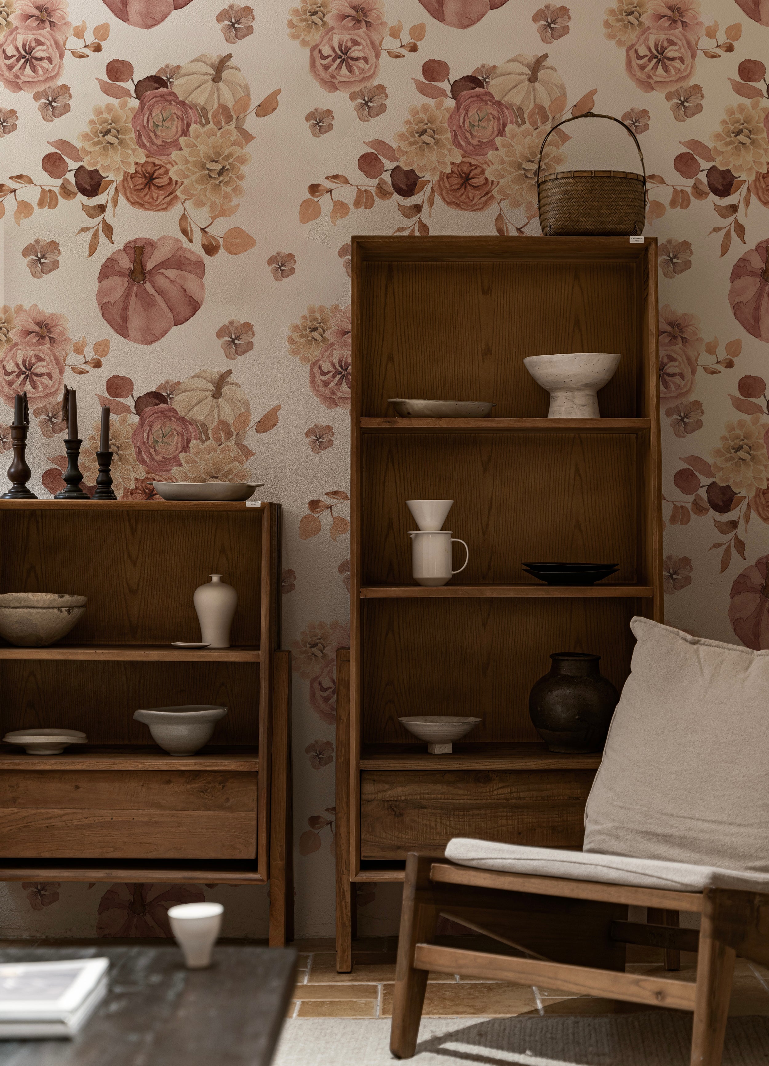 A cozy interior setting with a rustic wooden bookshelf filled with assorted pottery and candles against a wall adorned with Pumpkin Spice Wallpaper. The wallpaper features a lush, watercolor floral pattern in warm tones of pink, beige, and brown, with prominent pumpkin and flower motifs enhancing a vintage charm.