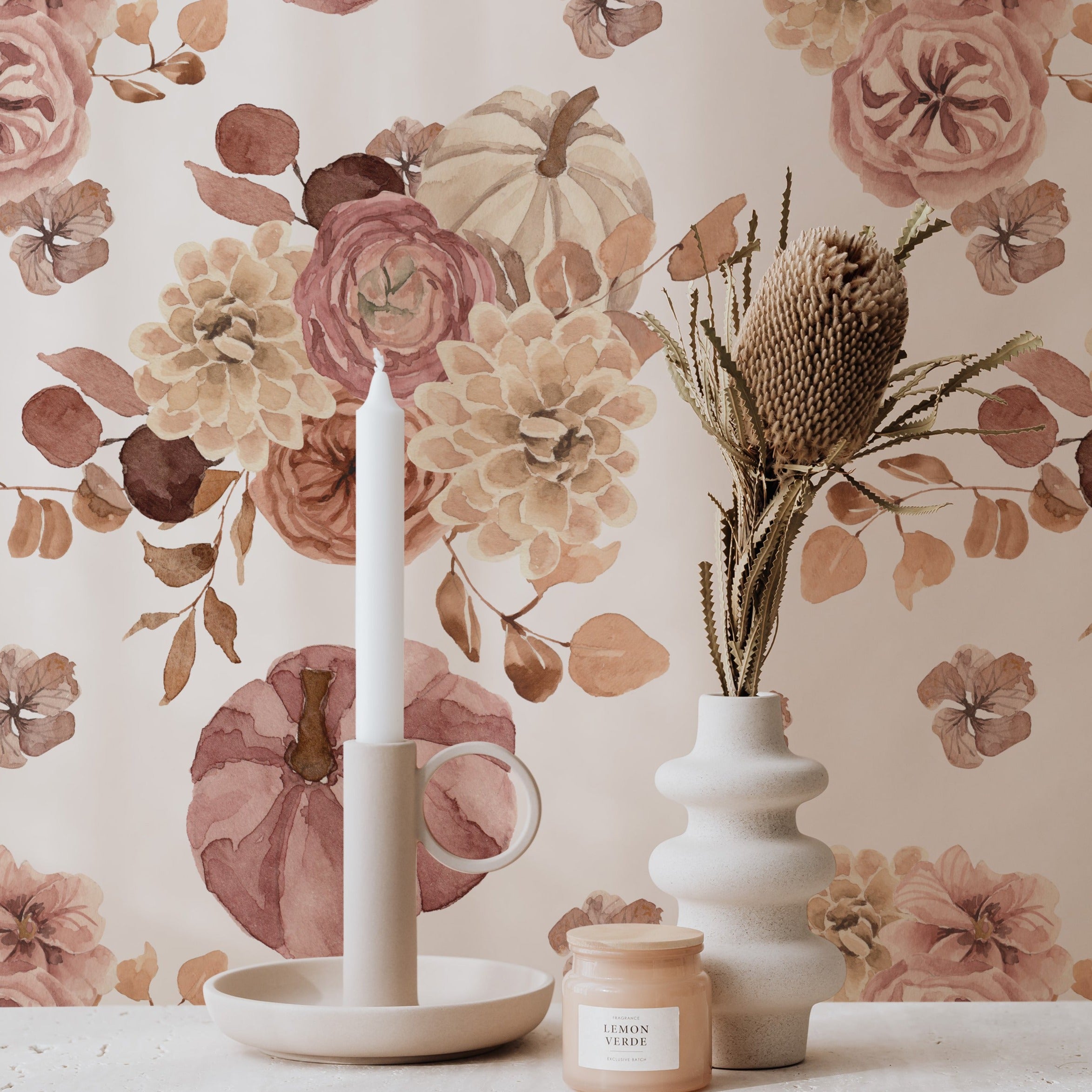 A well-styled corner of a room featuring the Pumpkin Spice Wallpaper as a backdrop. A simple modern decor includes a white candle in a tall ceramic holder, a dish with a candle, and a vase with dried botanicals, all set against the floral wallpaper that exudes a warm, inviting autumn vibe.