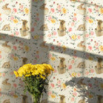 A bright room illuminated by sunlight, decorated with Garden Bunnies Wallpaper. The wallpaper's soft colors and playful rabbit motifs create a cheerful atmosphere, complemented by a bouquet of yellow flowers on a wicker table.