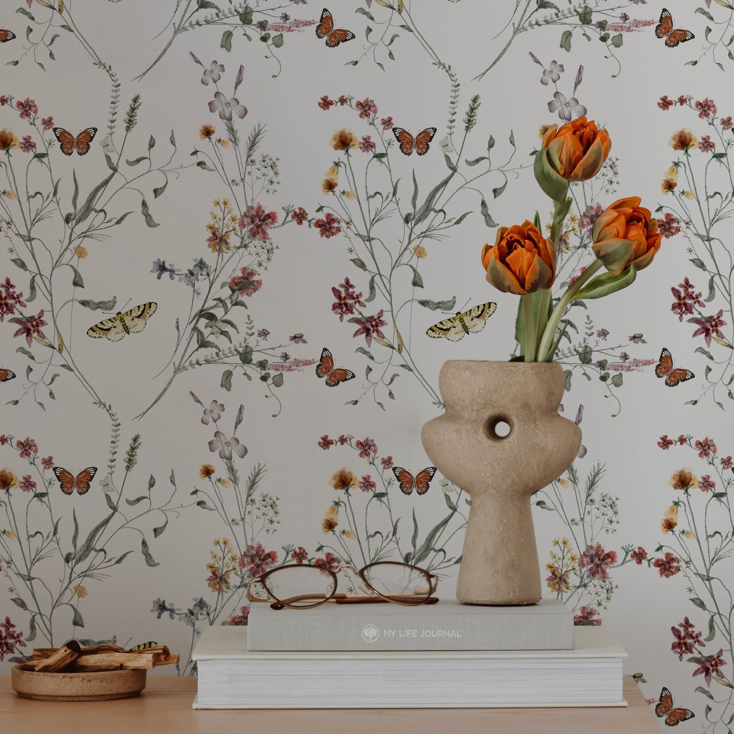 Stylish workspace with Butterfly Botanicals wallpaper. A ceramic vase with orange tulips, glasses, and books on a white table complement the floral theme
