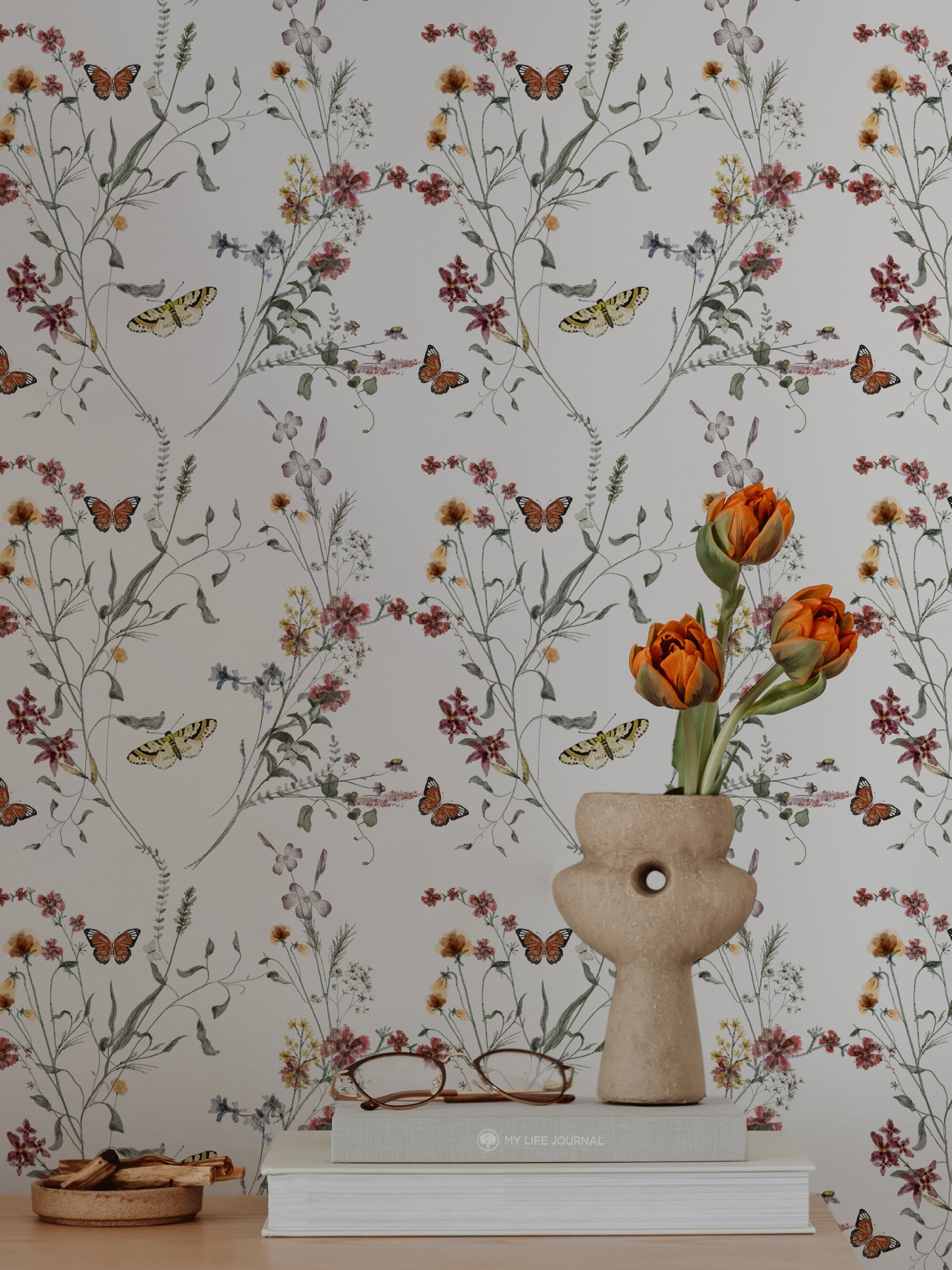 Stylish workspace with Butterfly Botanicals wallpaper. A ceramic vase with orange tulips, glasses, and books on a white table complement the floral theme