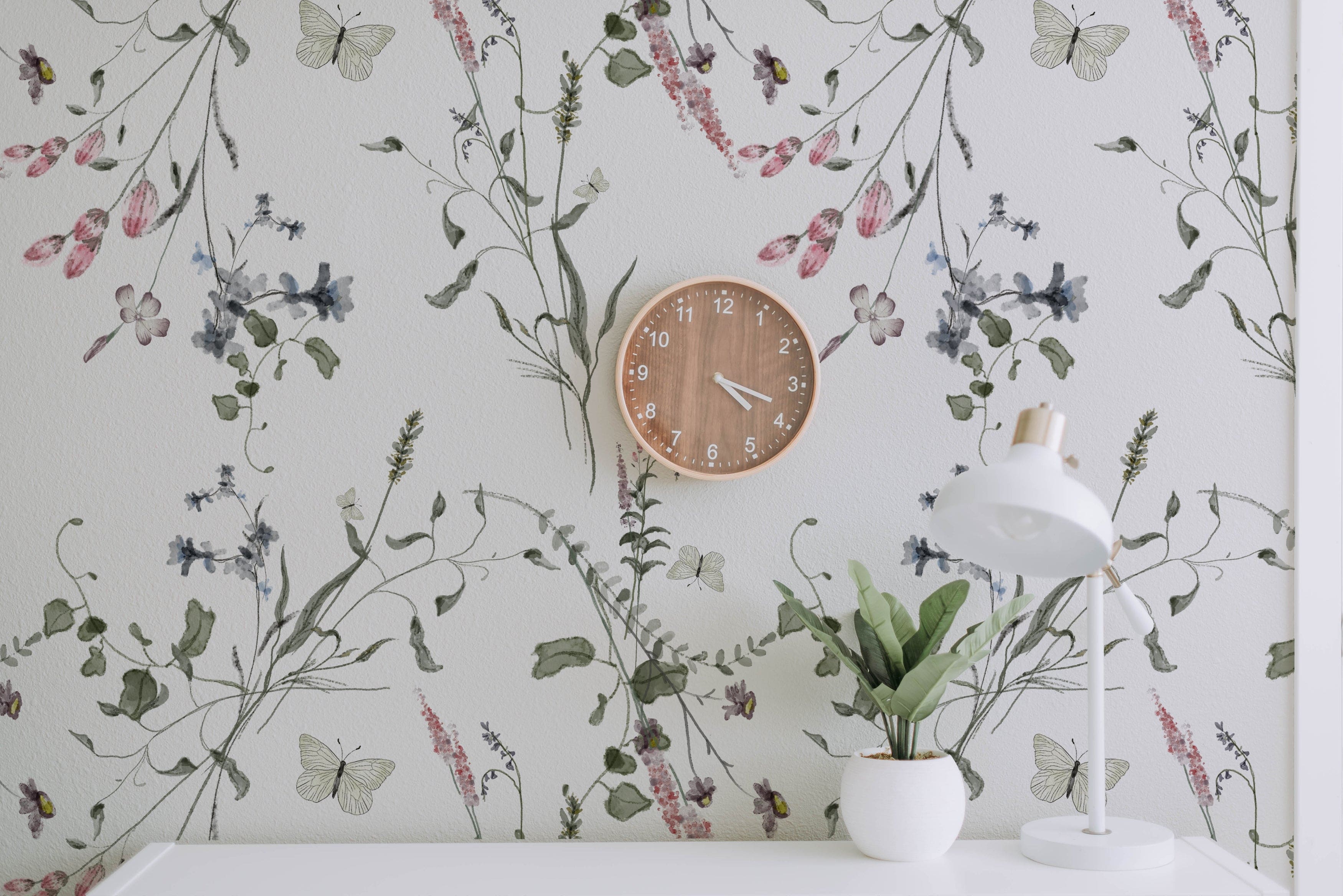 A serene office space enhanced by the Lovely Botanicals Wallpaper, featuring delicate botanical illustrations with flowers and butterflies in soft pastel colors. A wooden clock and white lamp on a sleek desk complement the gentle nature theme.