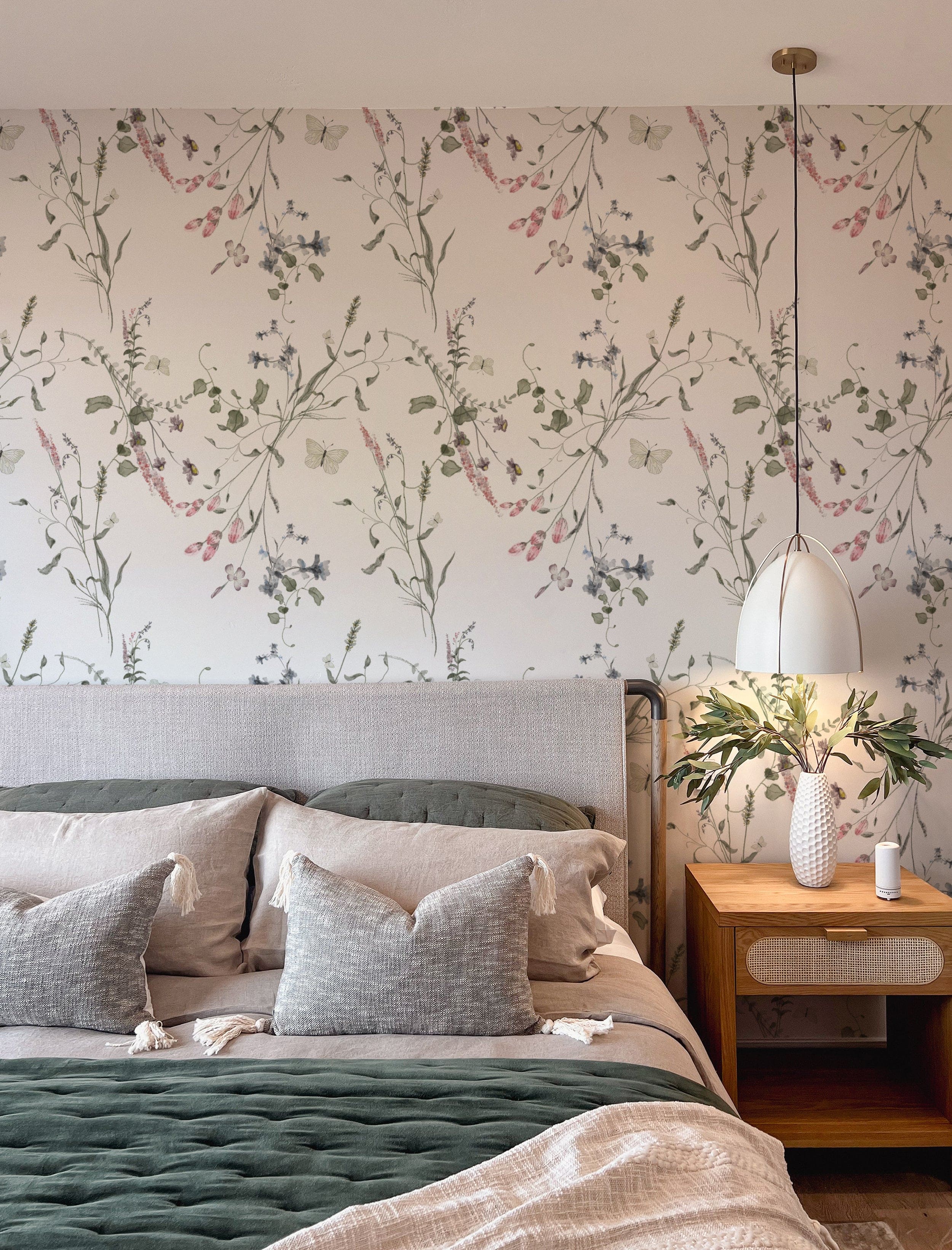 A cozy bedroom setting with the Lovely Botanicals Wallpaper creating a tranquil backdrop. The wallpaper's subtle floral and butterfly designs in soft hues add a soothing touch to the room, accompanied by plush pillows and a stylish bedside lamp.