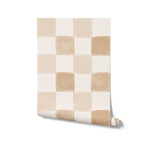 A roll of Clémence Wallpaper in Butterscotch, partially unrolled to showcase the checkered pattern of butterscotch and off-white squares. The soft, watercolor effect of the wallpaper provides a gentle and warm aesthetic, suitable for various interior design needs