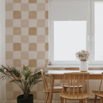 A bright dining area featuring Clémence Wallpaper in Butterscotch, characterized by large, square blocks in alternating shades of butterscotch and off-white, giving the space a warm and inviting atmosphere. The room is furnished with a simple wooden dining set and is decorated with a potted palm and a vase of dried flowers on the table.